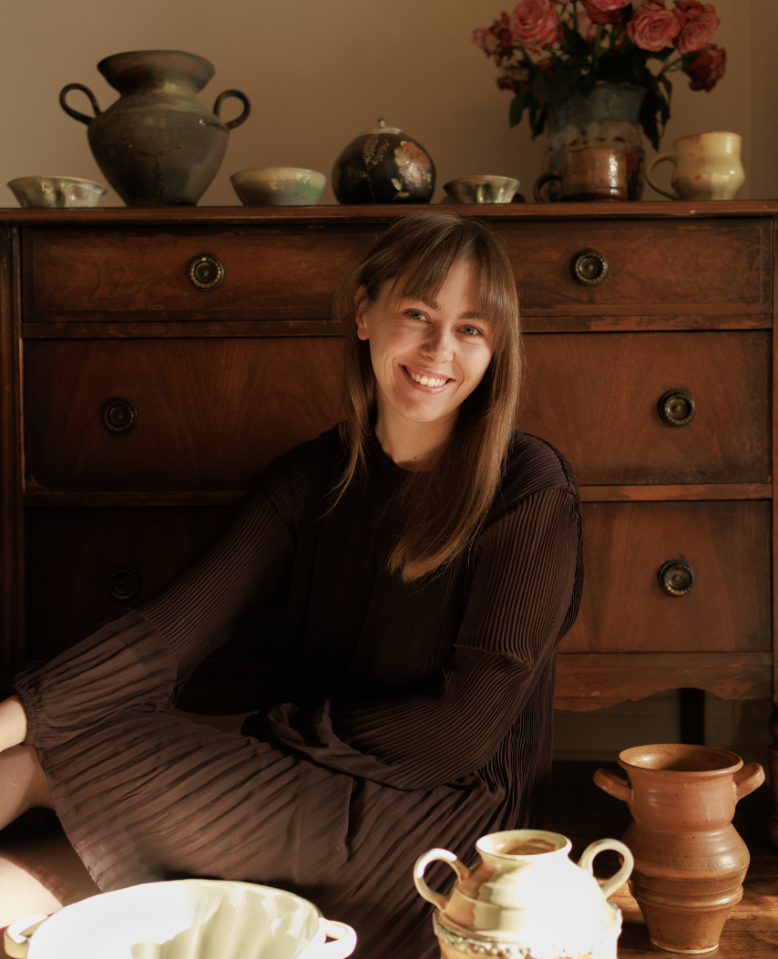 A young woman with long brown hair smiling while sitting on the floor in front of a wooden dresser with decorative pottery and a flower arrangement on top.