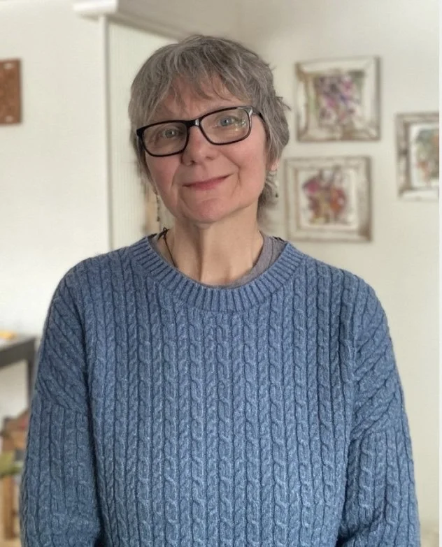 A woman with short gray hair and glasses wearing a blue cable-knit sweater, standing indoors with framed pictures on the wall behind her.
