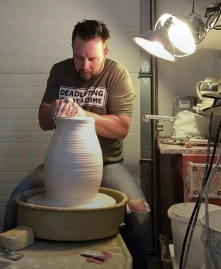 Man shaping a tall, white ceramic vase on a pottery wheel in a pottery studio.