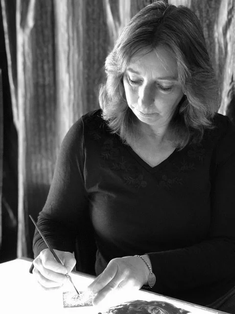 A woman with shoulder-length hair in a black long-sleeve shirt, sitting at a table, writing or drawing with a brush on a piece of paper or canvas in a room with curtains or drapes in the background.