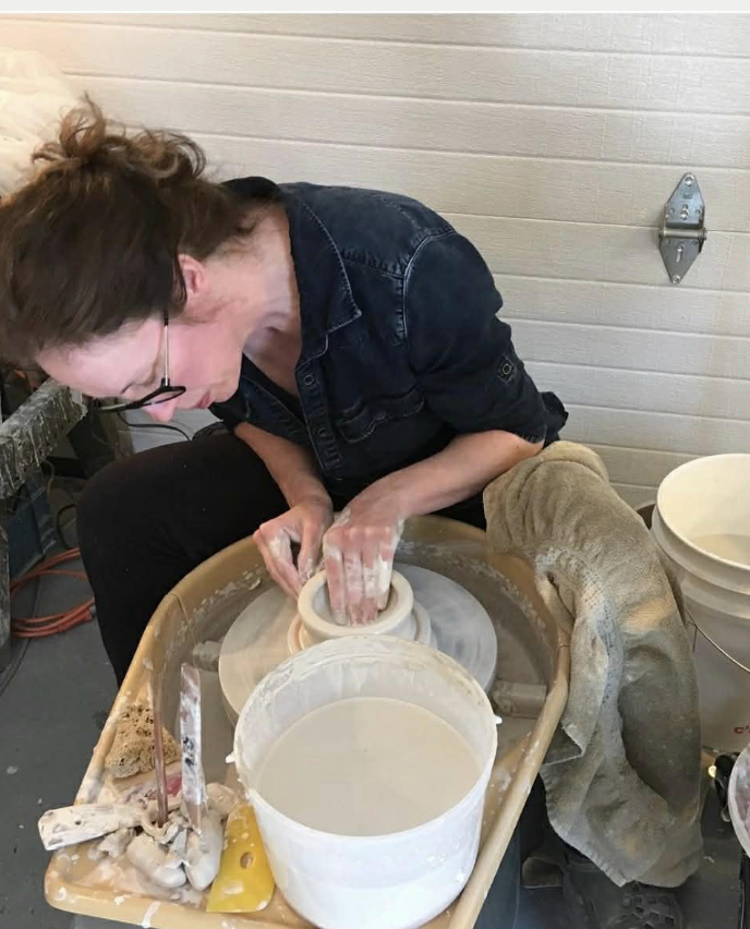 A woman shaping a clay pot on a pottery wheel in a workshop.