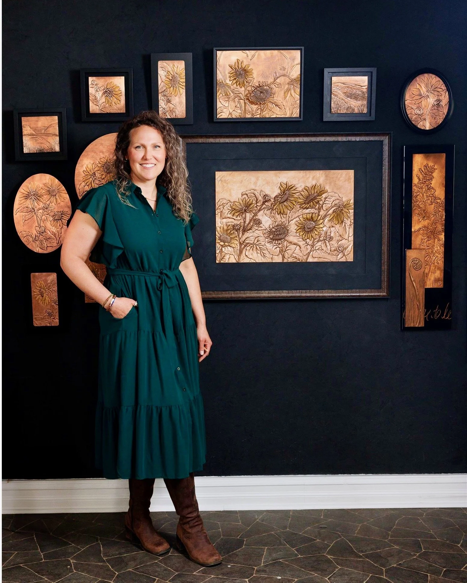 Erin Stelmaschuk Biegel, southern Alberta, copper artist. Standing in front of a black wall decorated with framed botanical artwork featuring handmade copper artwork of flowers, prairie scenes and landscapes.