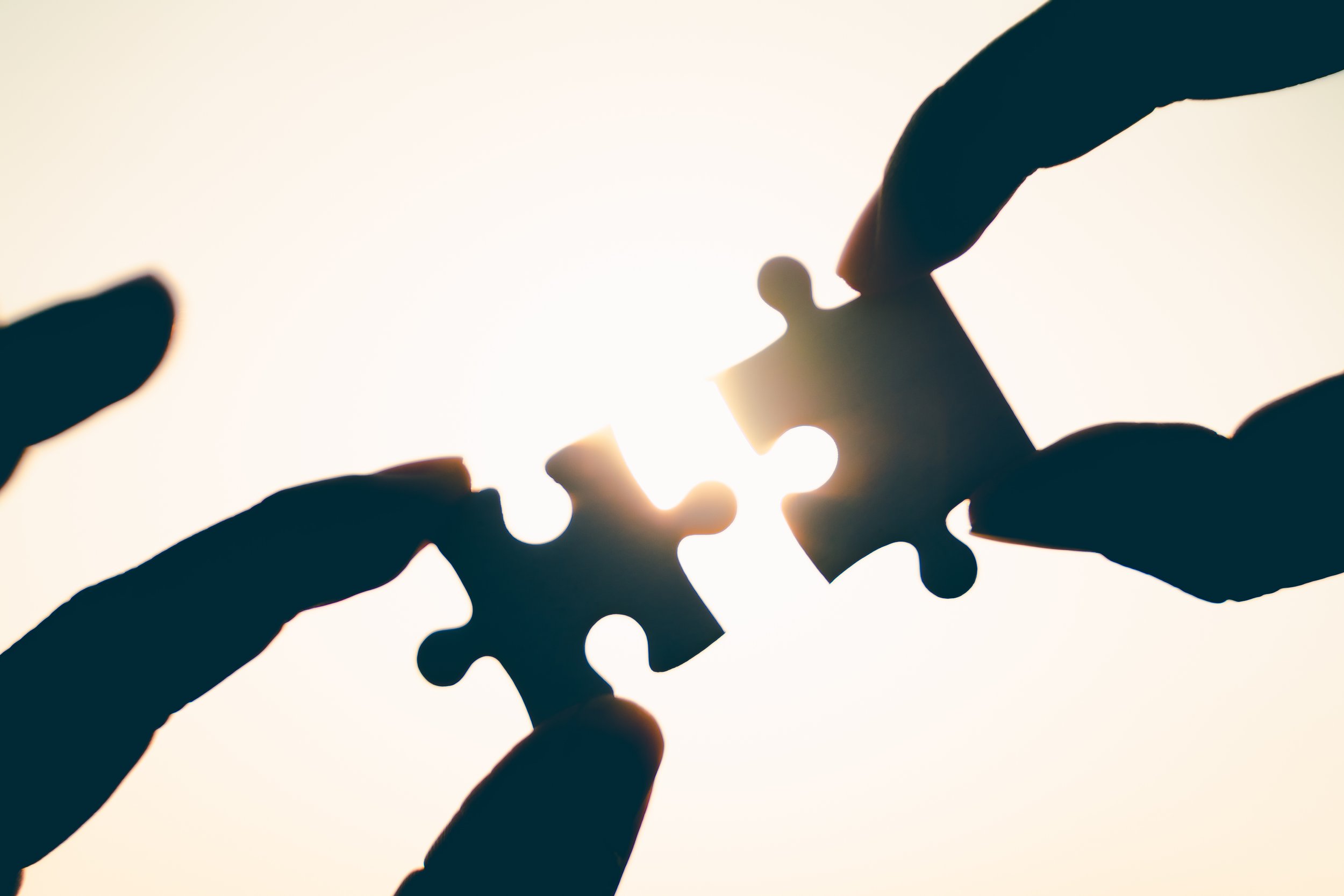 Two silhouetted hands holding puzzle pieces connecting against a bright background.
