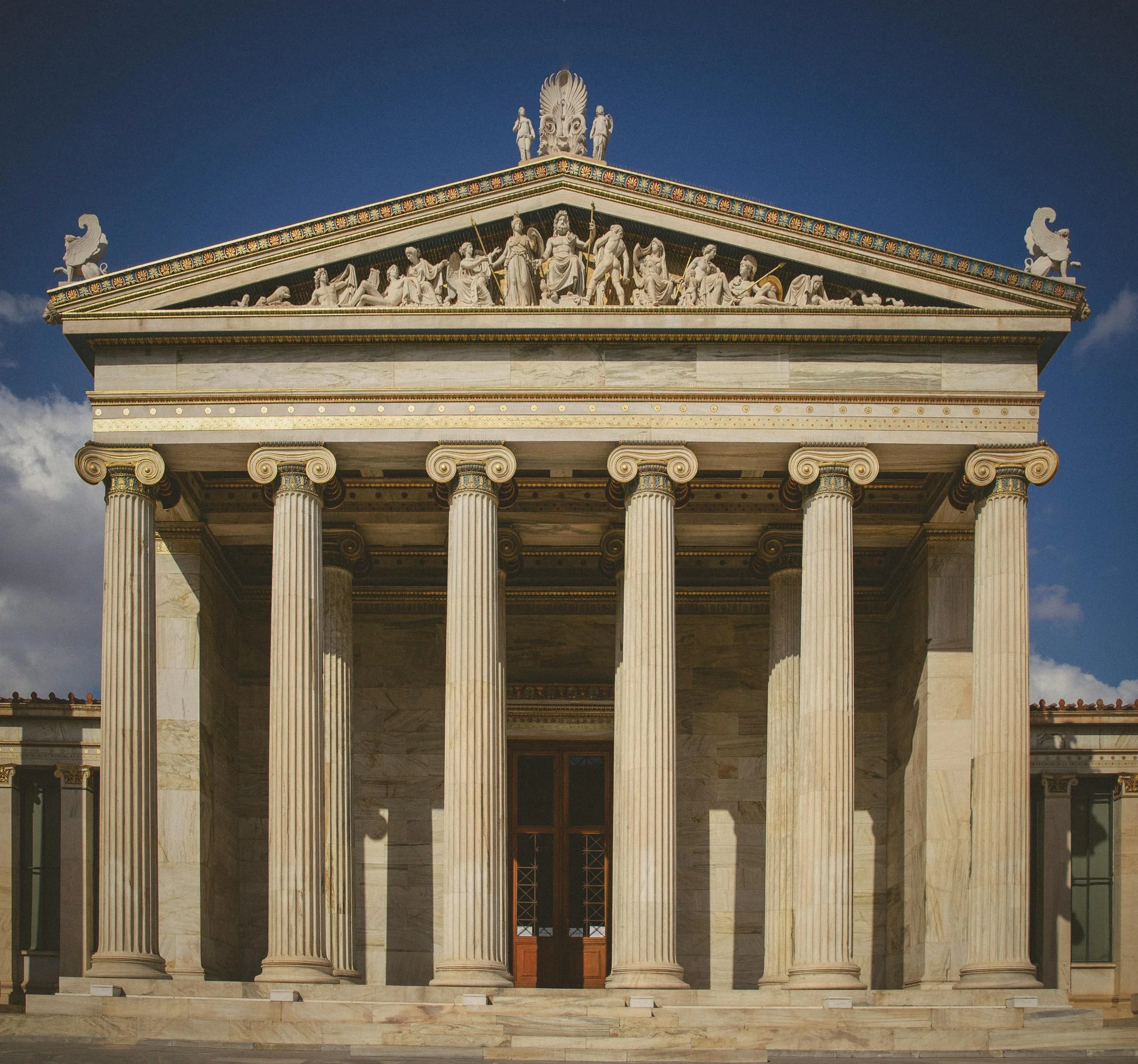 Front view of the Greek Parthenon temple with six Corinthian columns and a detailed sculptural frieze on the pediment, set against a blue sky.