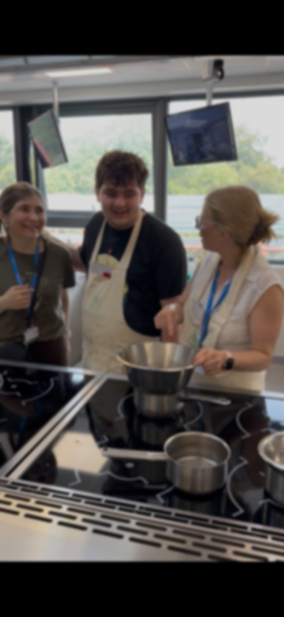 Three people in a kitchen, wearing aprons, standing around a stove with saucepans, smiling and engaging with each other.