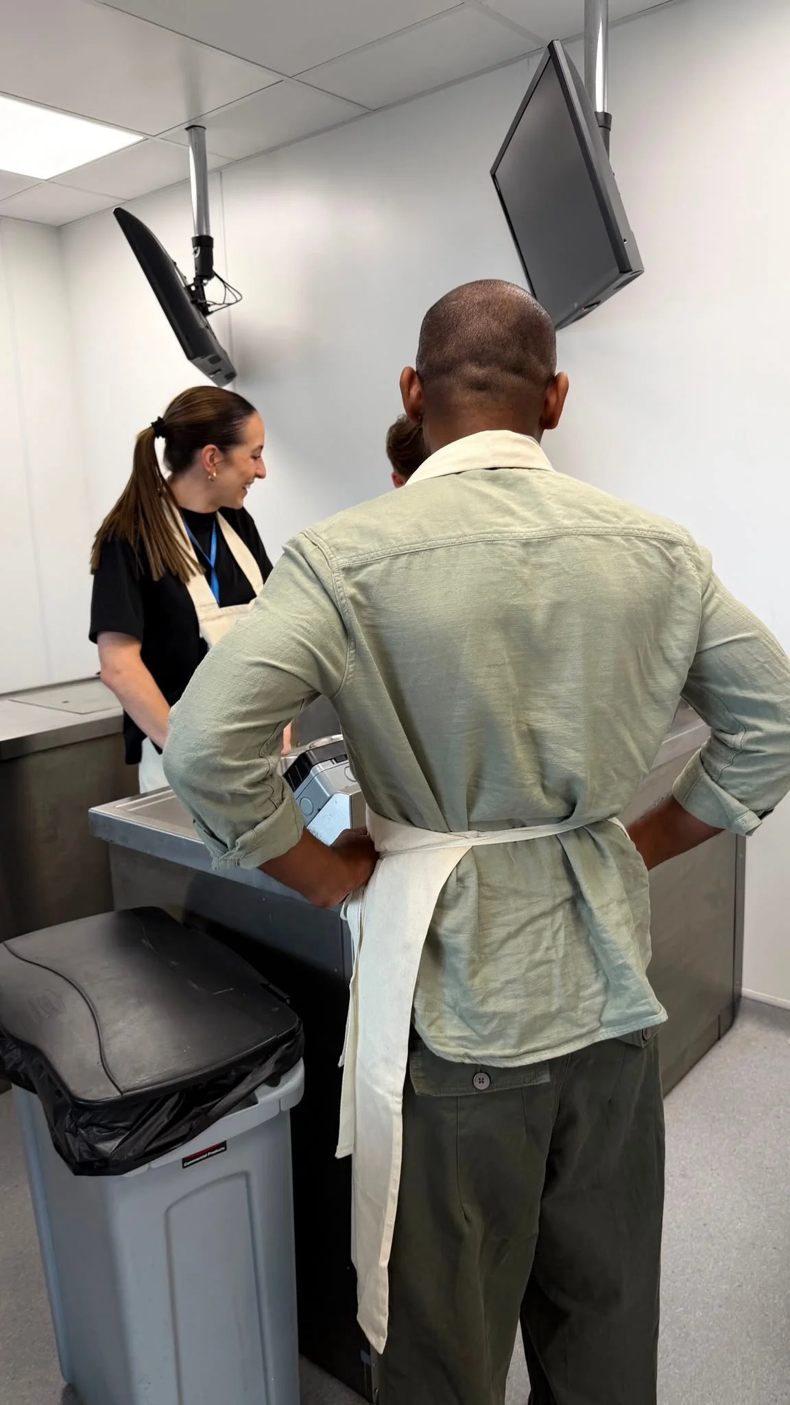 A man in green uniform with a white apron stands with hands on hips, facing a woman with long brown hair in a black shirt, in a room with white walls and a ceiling-mounted TV and speakers.