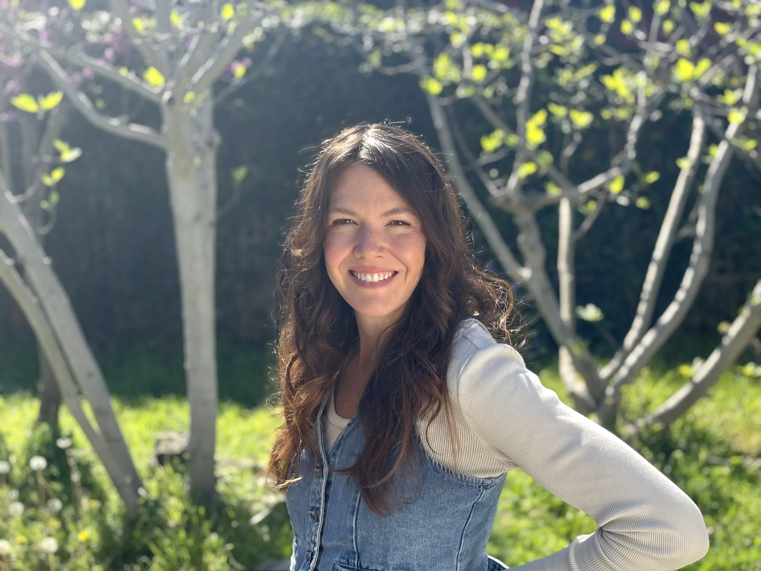 A woman smiling outdoors in sunlight, standing in front of trees with green leaves.