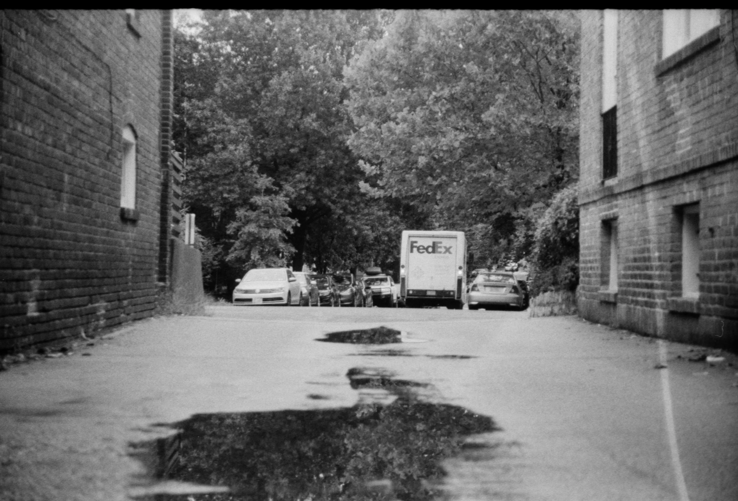 Black and white photo of a street alley with puddles, parked cars, trees in the background, brick buildings on either side, and a FedEx truck.