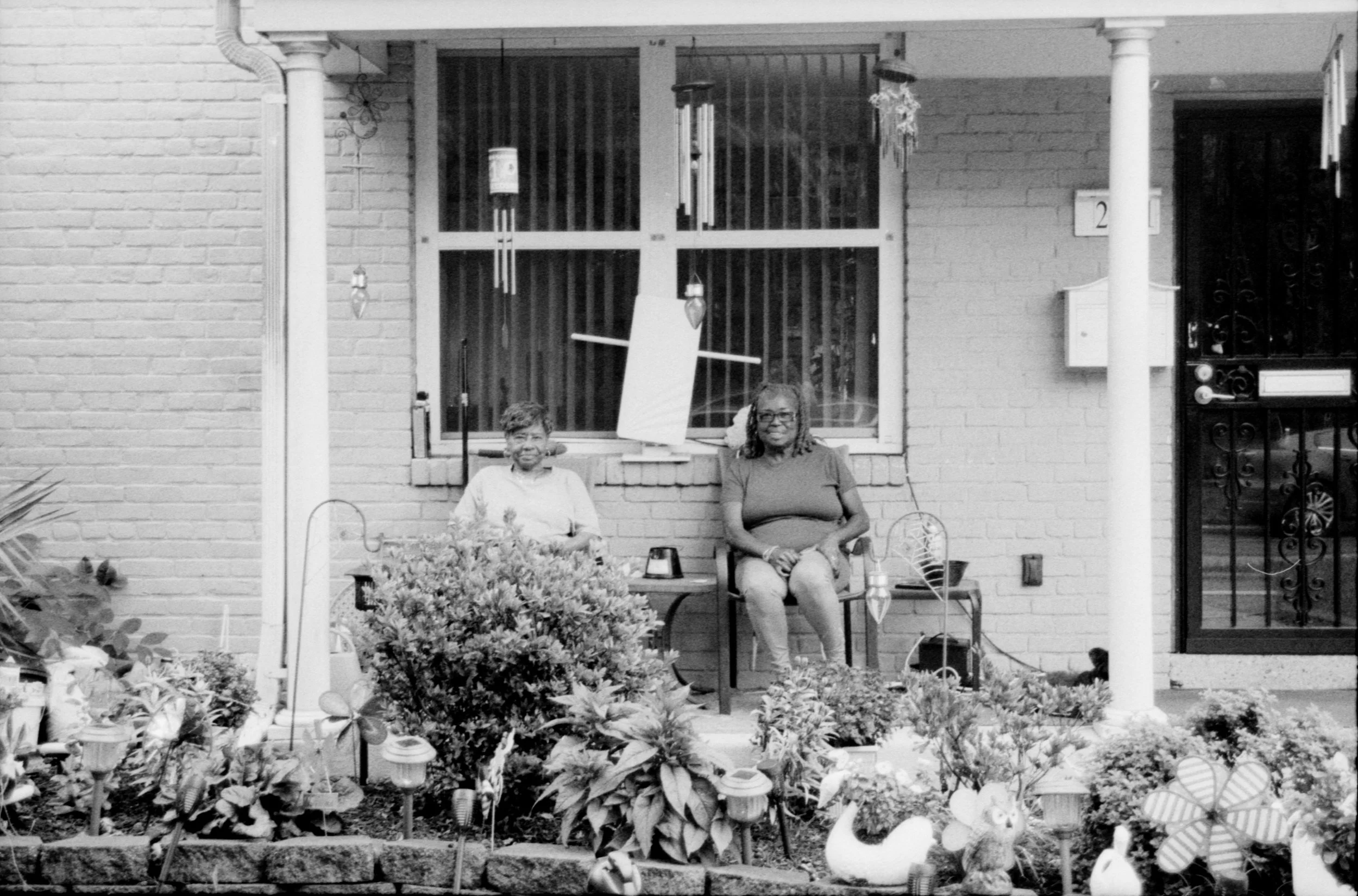 Black and white photo of two women sitting on a porch, surrounded by plants and garden decorations.