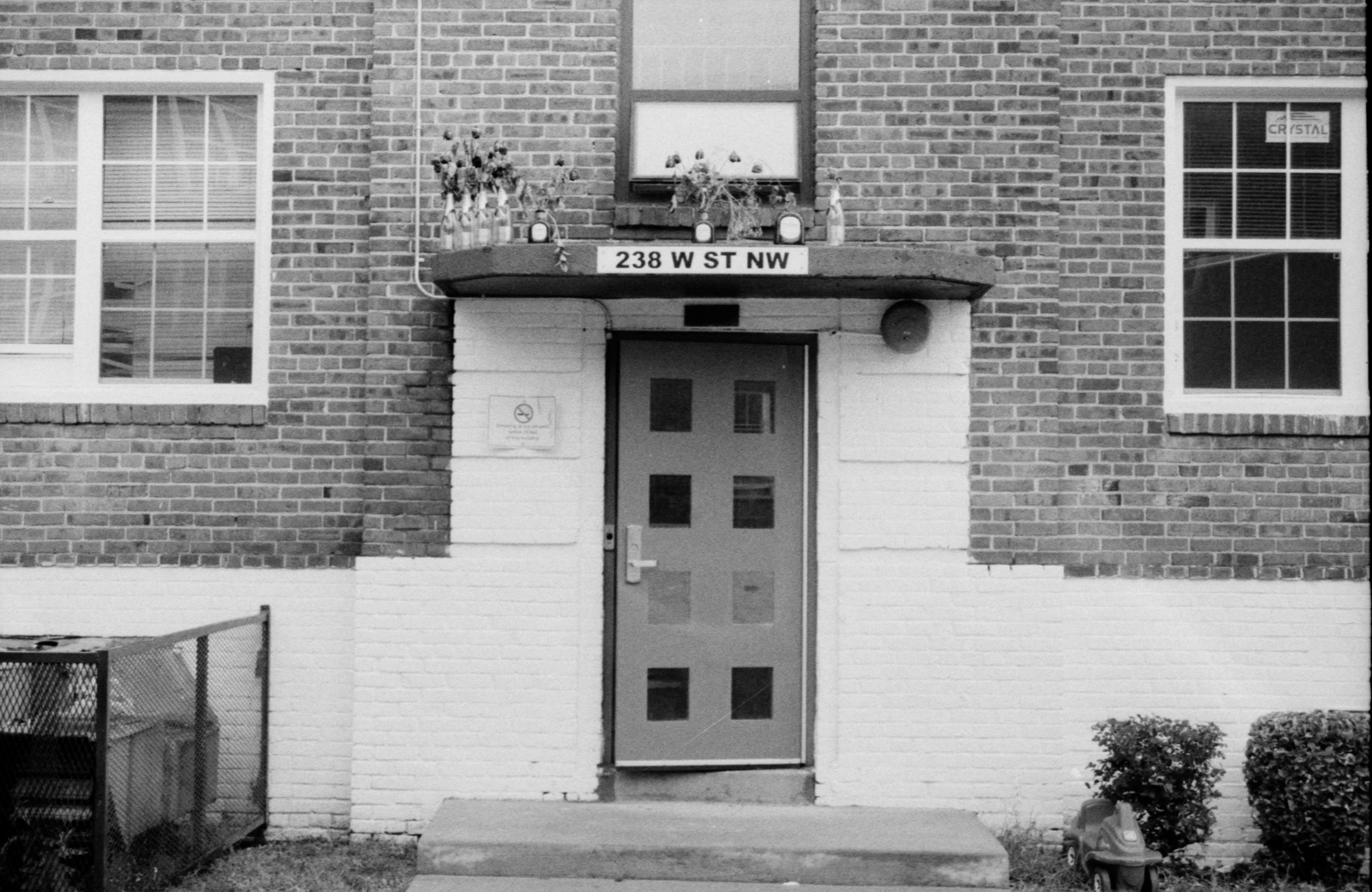 Black and white photo of apartment building entrance with a door, two windows, and a sign that reads '238 W ST NW.' There are potted plants and flowers on a small balcony above the door, and a no smoking sign on the wall next to the door. Bushes and 