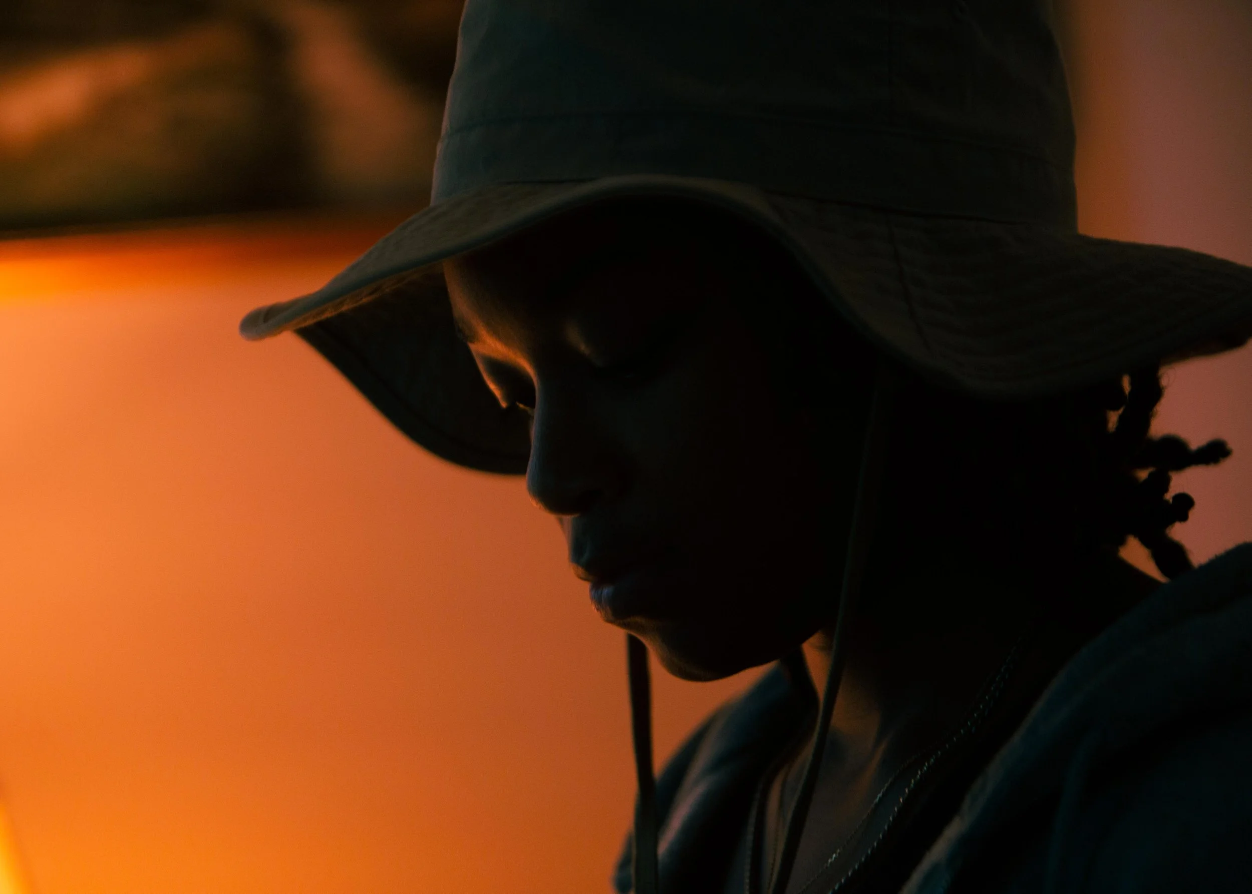 A person wearing a bucket hat, head bowed, with sunset in the background.