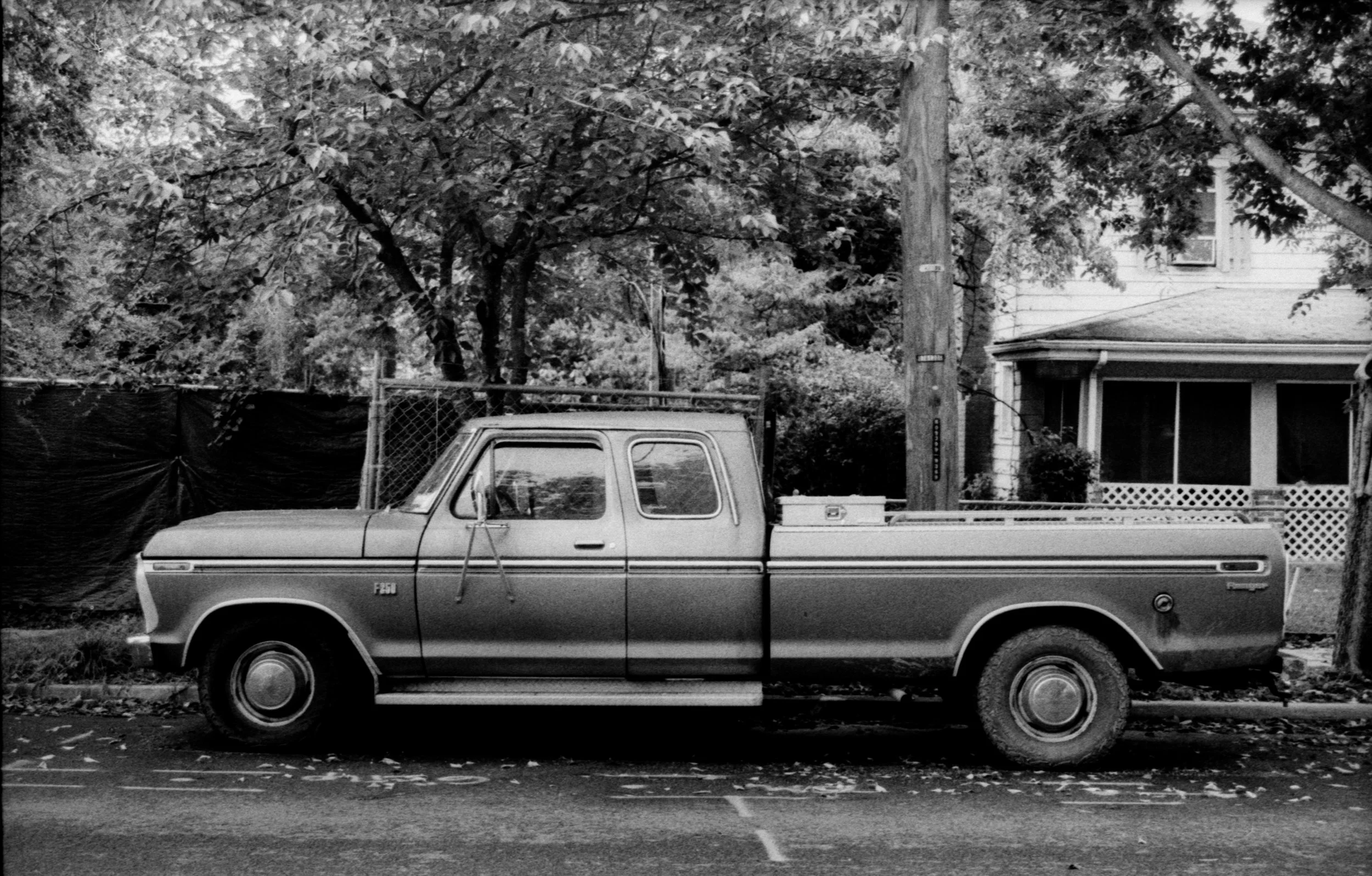 Black and white photo of an old pickup truck parked on a residential street with trees and a house in the background.