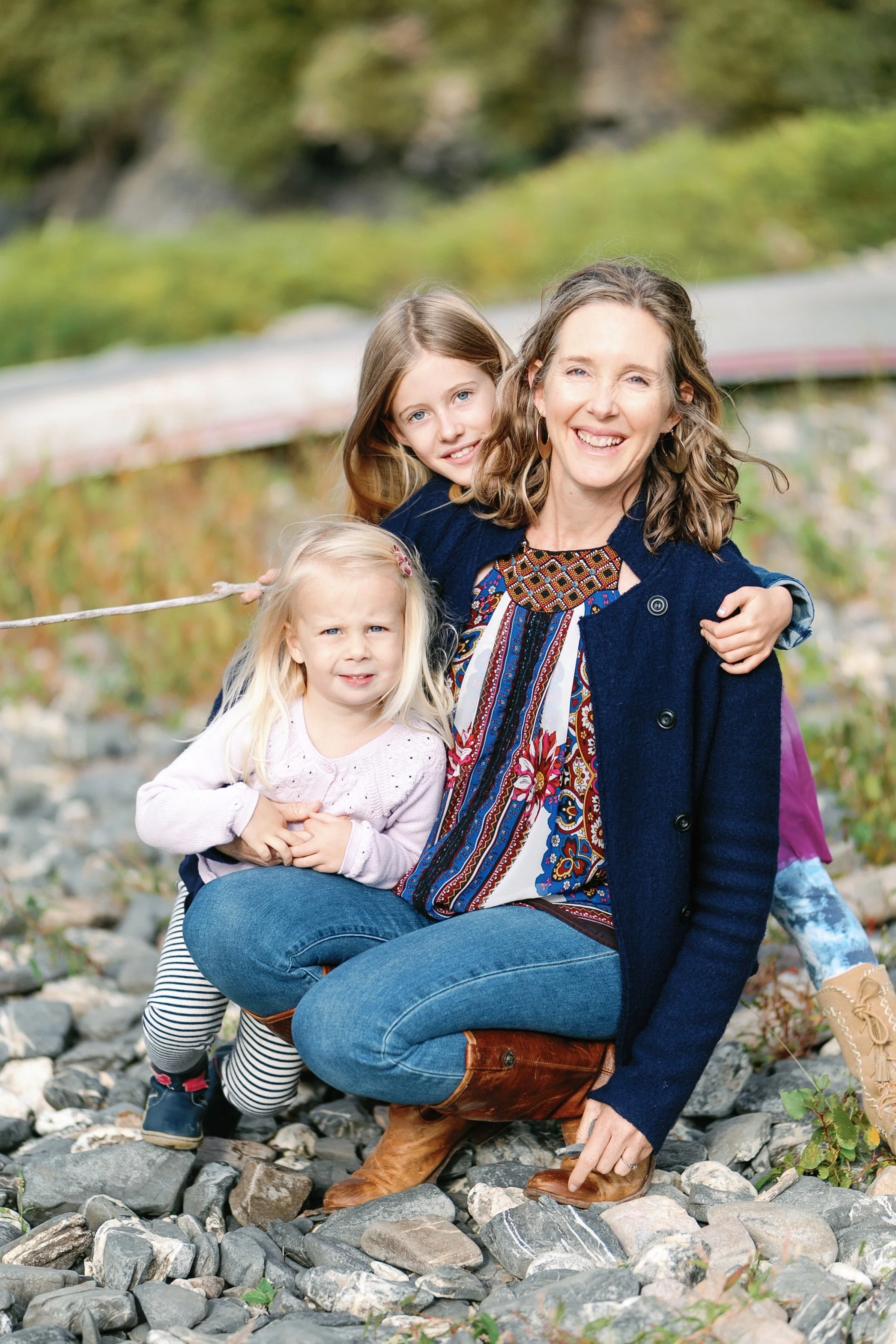 A woman with two young girls outdoors on a rocky surface, smiling. The woman is wearing a patterned blouse, dark coat, and jeans. The girl behind her has brown hair, and the girl sitting on her lap has blonde hair, with a worried expression.
