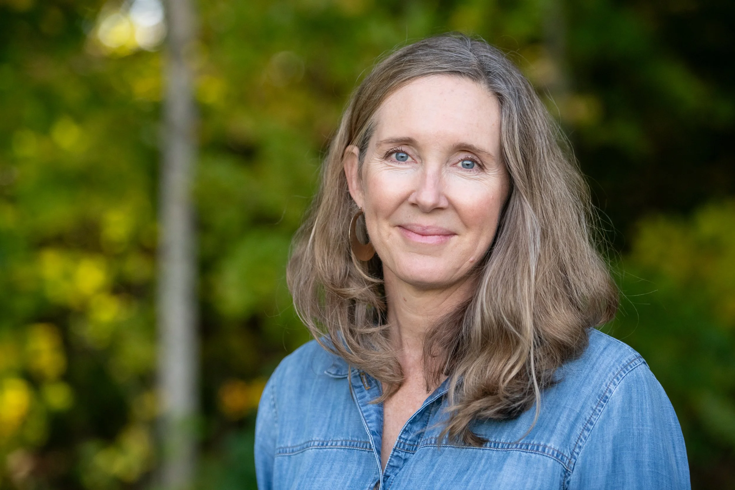 A woman with long, wavy, light brown hair and blue eyes, smiling softly, wearing a denim shirt and hoop earrings, outdoors with a blurry background of green trees.