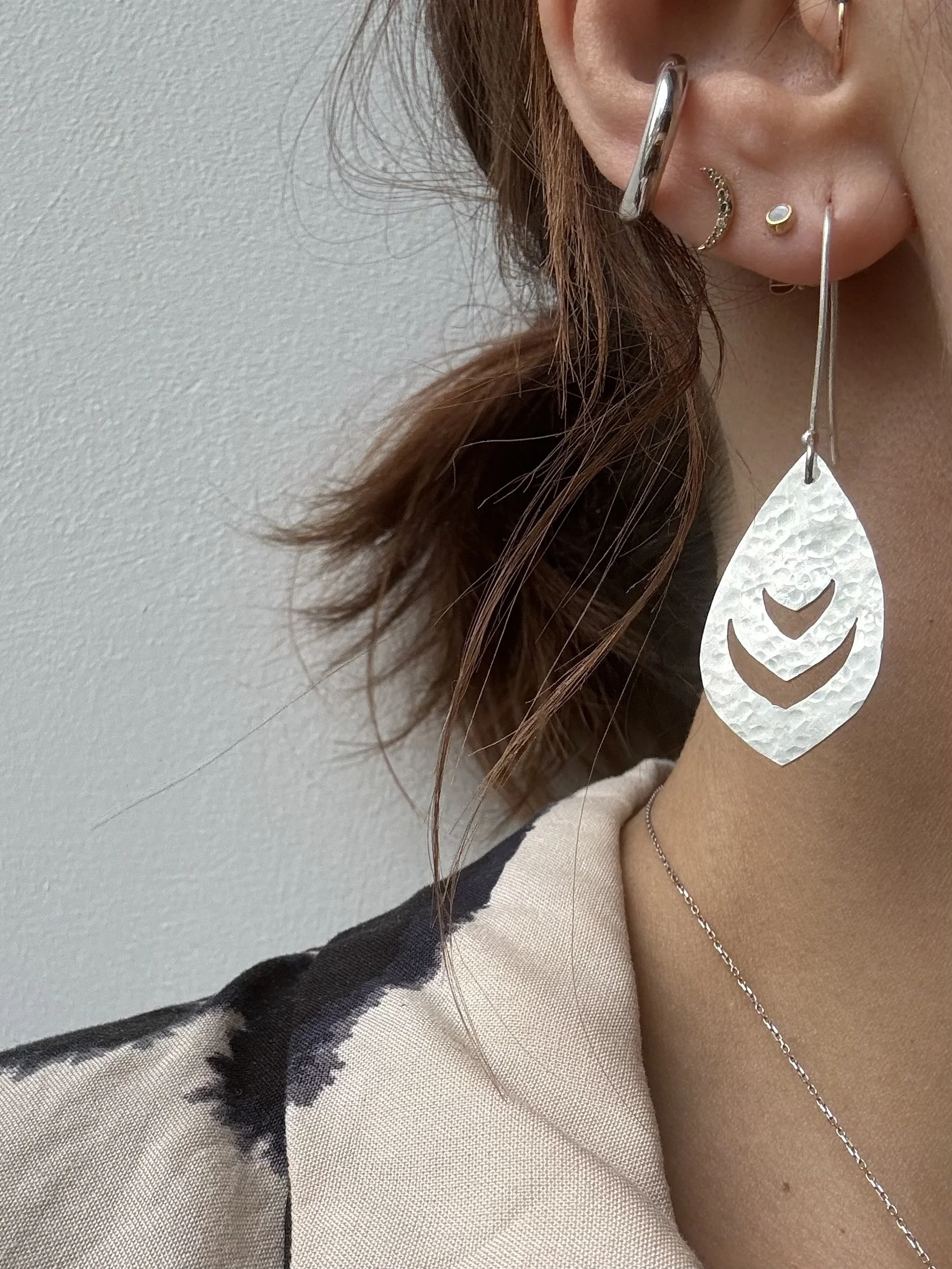 Close-up of a woman's ear with multiple earrings, a large silver earring, a small hoop with decorative stones, a small round stud, and a long silver drop earring featuring a textured teardrop-shaped pendant.
