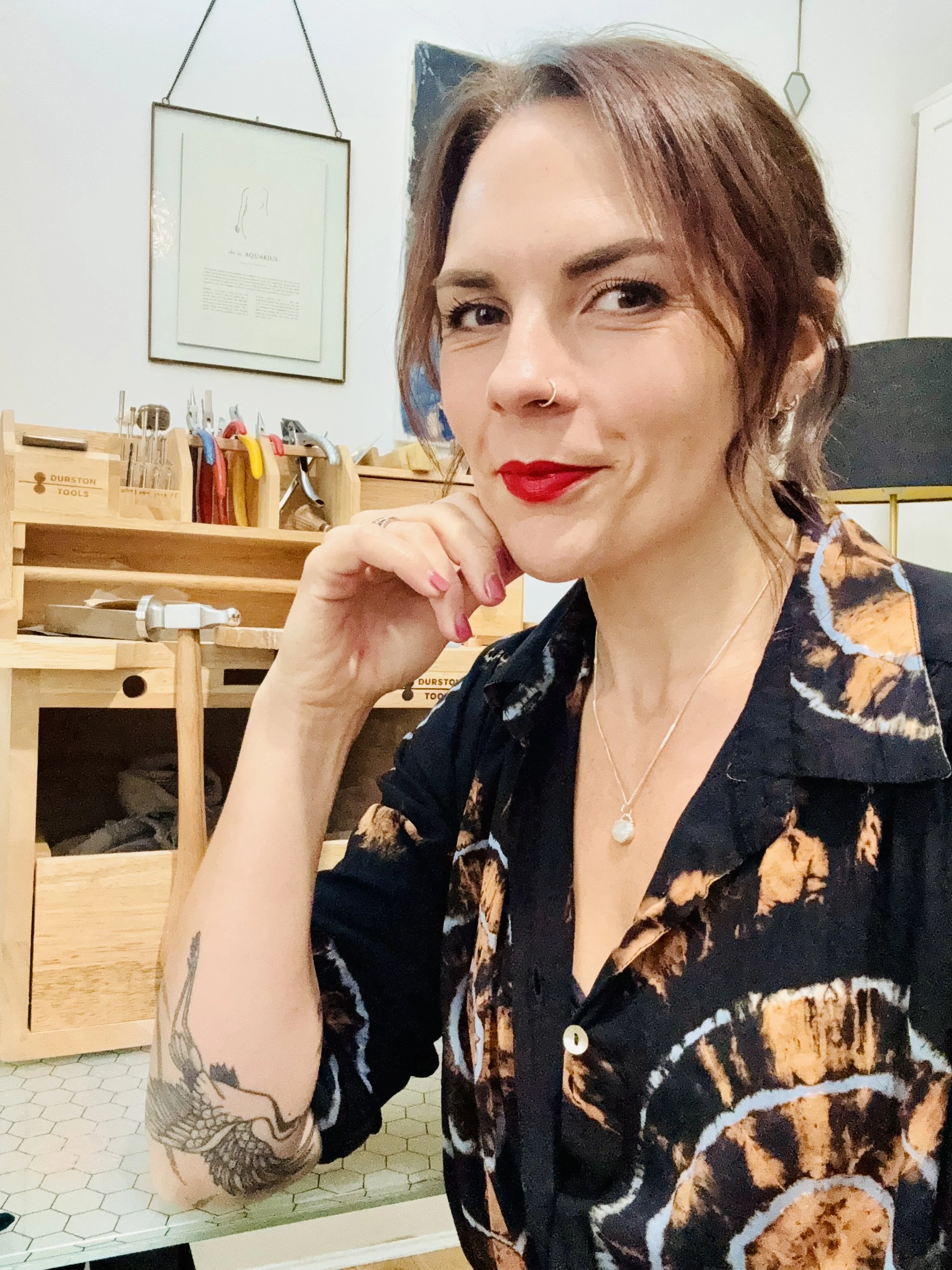 A woman with short, wavy brown hair posing with her hand near her chin, wearing a bold patterned shirt and a pearl necklace. She has a nose piercing, red lipstick, and is smiling slightly. Behind her, there is a wooden jewellery workbench with tools.