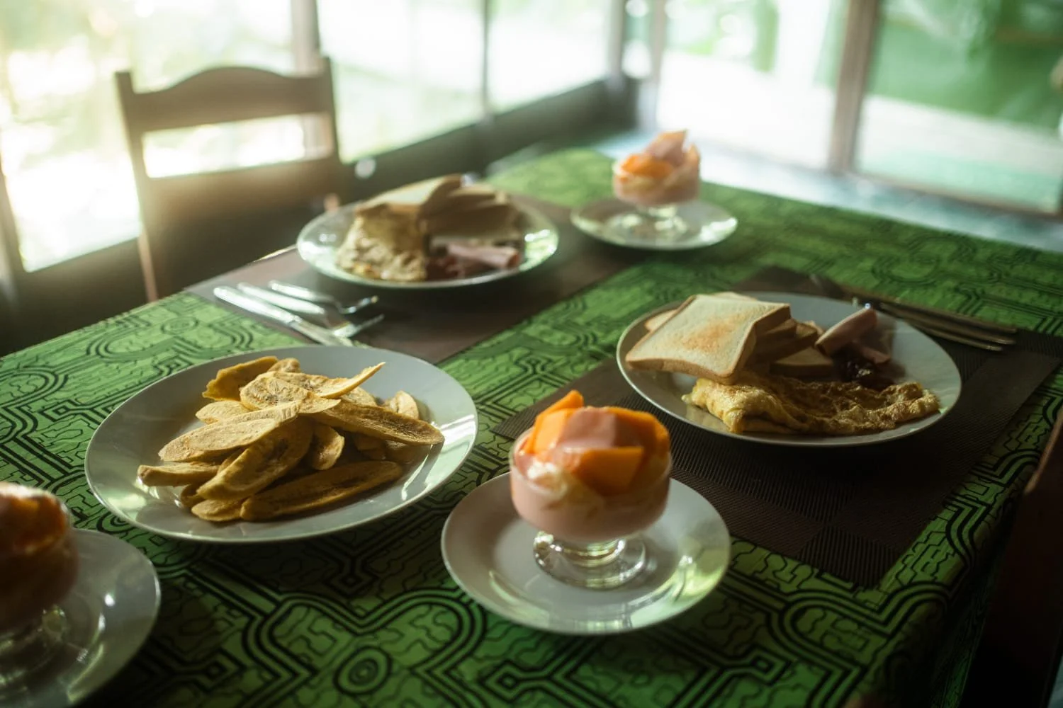 A table set with various breakfast foods, including toast, fried eggs, sliced meat, and fruit parfaits, on a green patterned tablecloth.