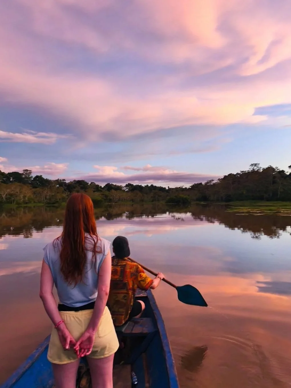 A woman with red hair stands on a boat looking at a person paddling on a river during sunset, with colorful clouds and trees reflected in the water.