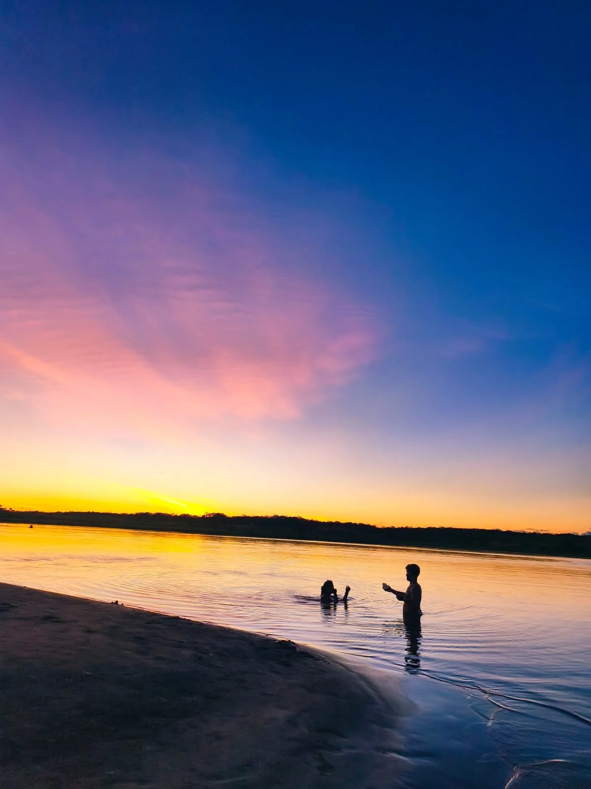 People enjoying a sunset at the beach, with three individuals in the water, one holding a phone, and the sky painted in pink, purple, and blue hues.