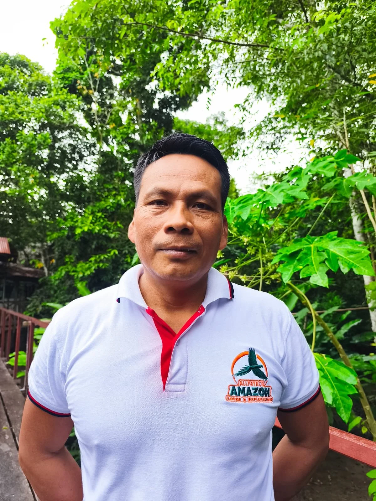 A man stands outdoors in front of lush green foliage, wearing a white polo shirt with a logo that reads 'Allpacayoc Amazon Lodge & Explorers'.