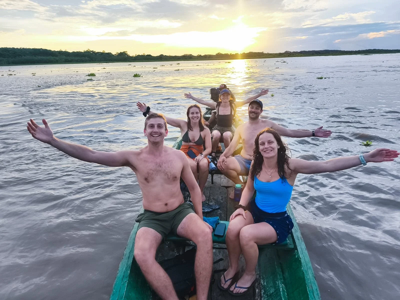Group of six friends sitting on a boat in the water at sunset, smiling and spreading their arms.