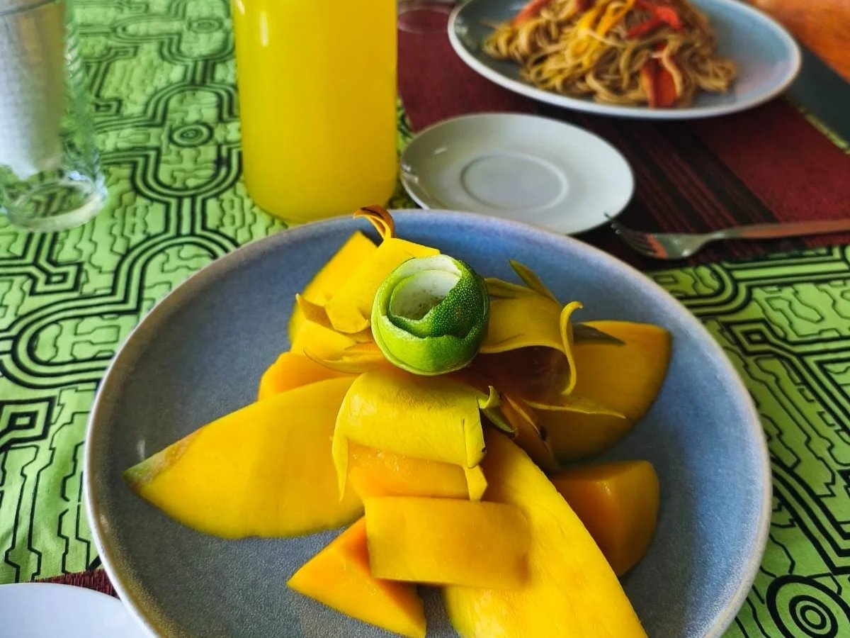 A plate of sliced mango with a small green lime on top, set on a vibrant green and black patterned tablecloth. In the background, there is a glass of water, a yellow bottle, a plate of noodles with vegetables, and an empty small white plate.