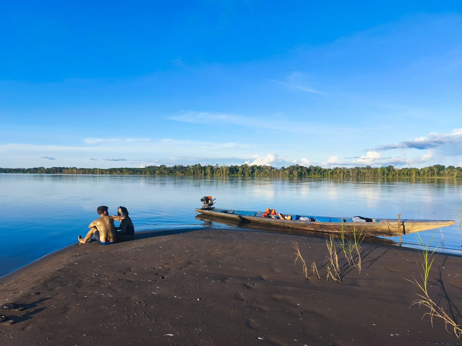 A couple sitting on a sandy riverbank near a boat, with calm water and a distant tree-lined shoreline under a blue sky with some clouds.