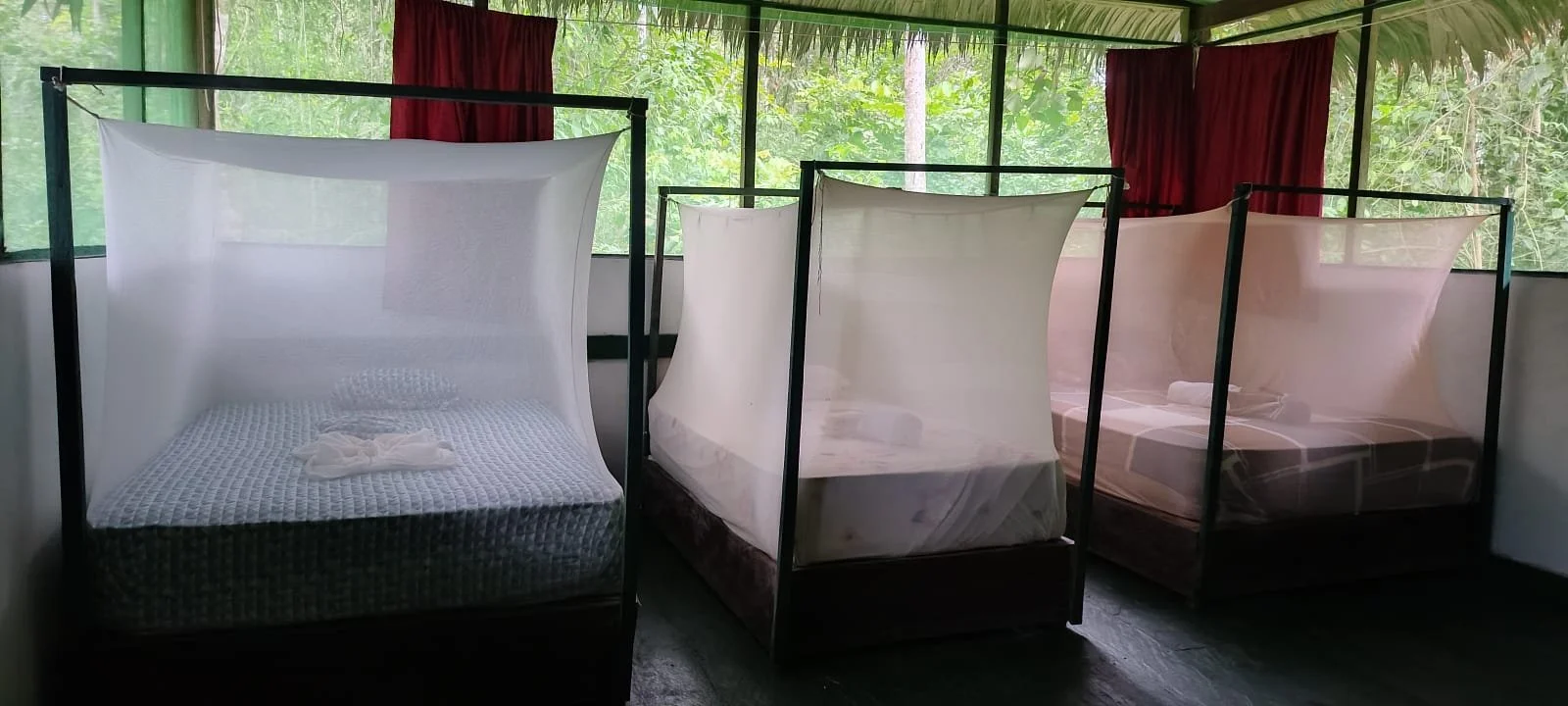 Three beds with mosquito nets in a room with green foliage view outside. The bed on the left has a towel folded on it. The middle bed is empty. The right bed has a pillow and small blanket. Red curtains are partially closed behind the beds.