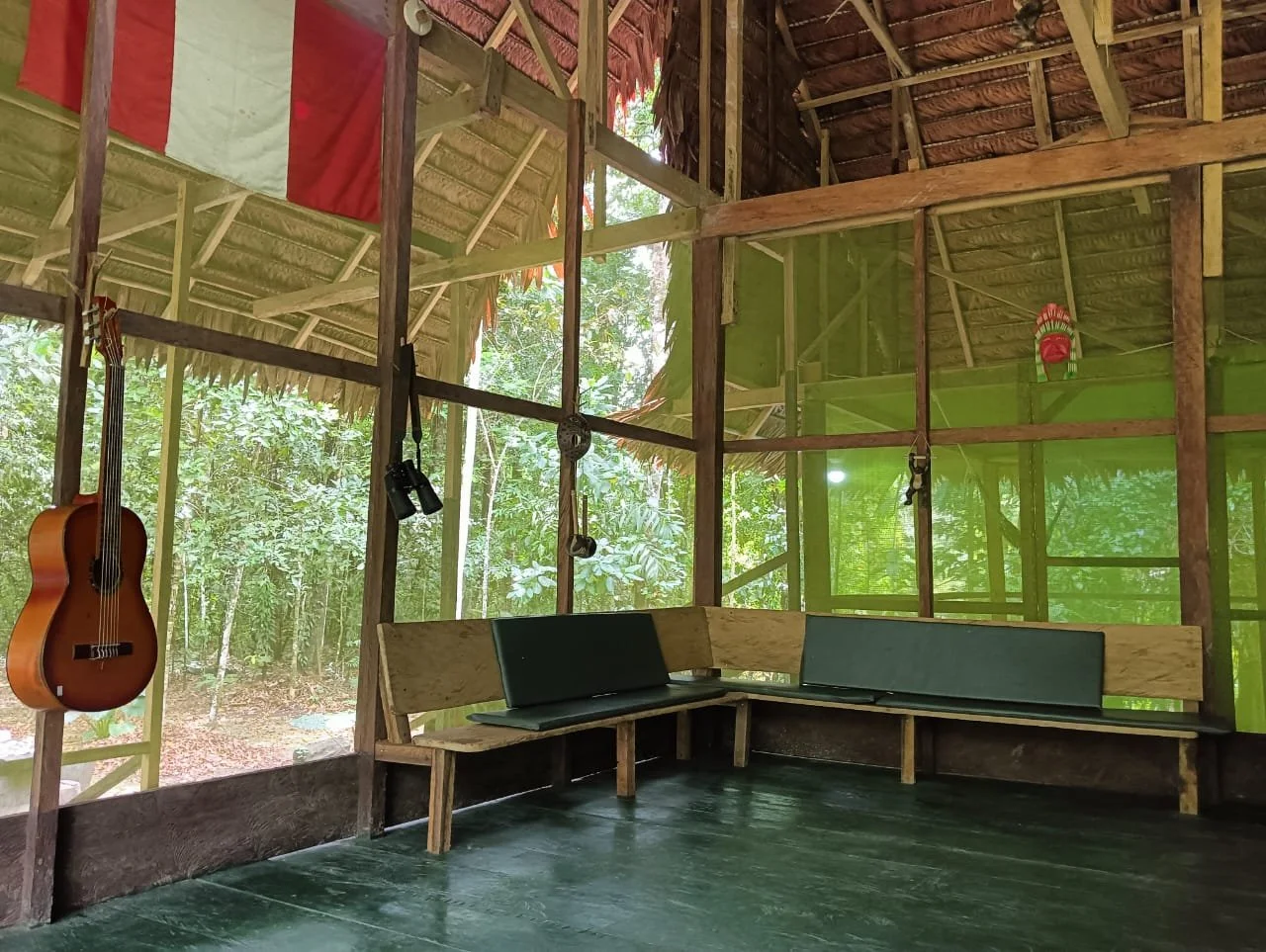 A rustic wooden room with a green netted wall, a wooden bench with dark cushions, a hanging guitar, and binoculars on hooks. The room opens to a lush outdoor jungle area.