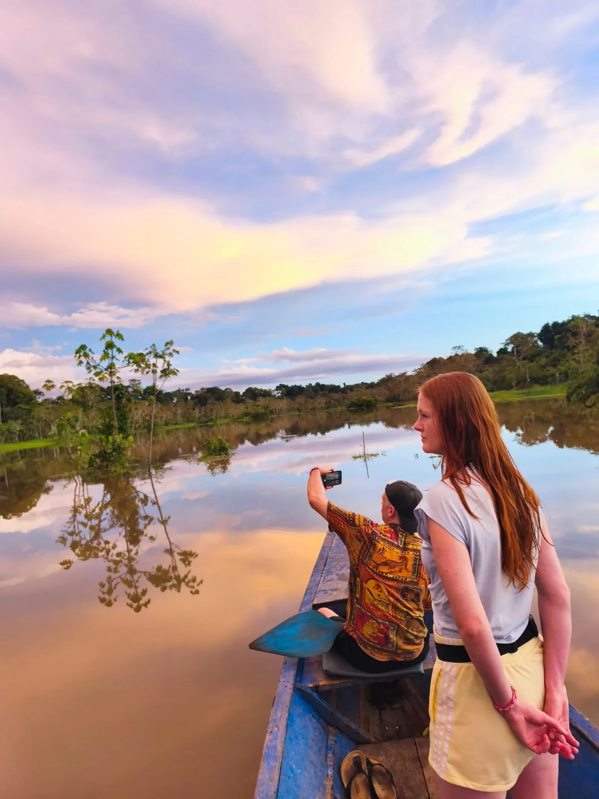 Two people on a boat taking a photo of a calm river with trees and a colorful sky reflected in the water.