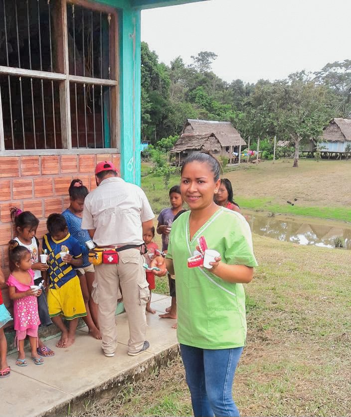 A young woman in a green shirt and jeans is smiling and holding small containers with white substance, likely ice cream. Behind her, a group of children and an older man are standing outside a rural building, some holding similar containers, with traditional huts and trees in the background.