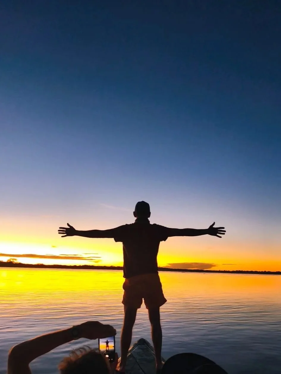 Person standing on a paddleboard with arms outstretched at sunset over a body of water, with another person taking a photo.