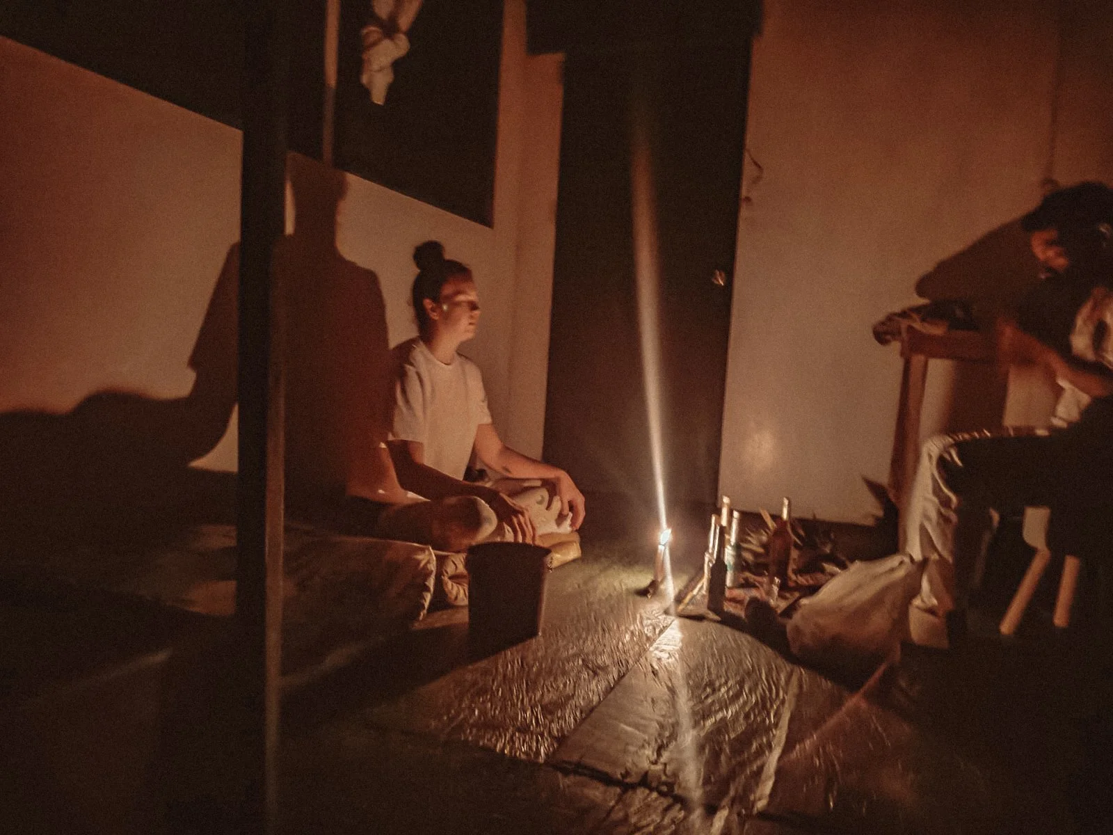 Woman sitting cross-legged on the floor in a dimly lit room lit by a single candle, with another person sitting nearby, and various objects scattered around.