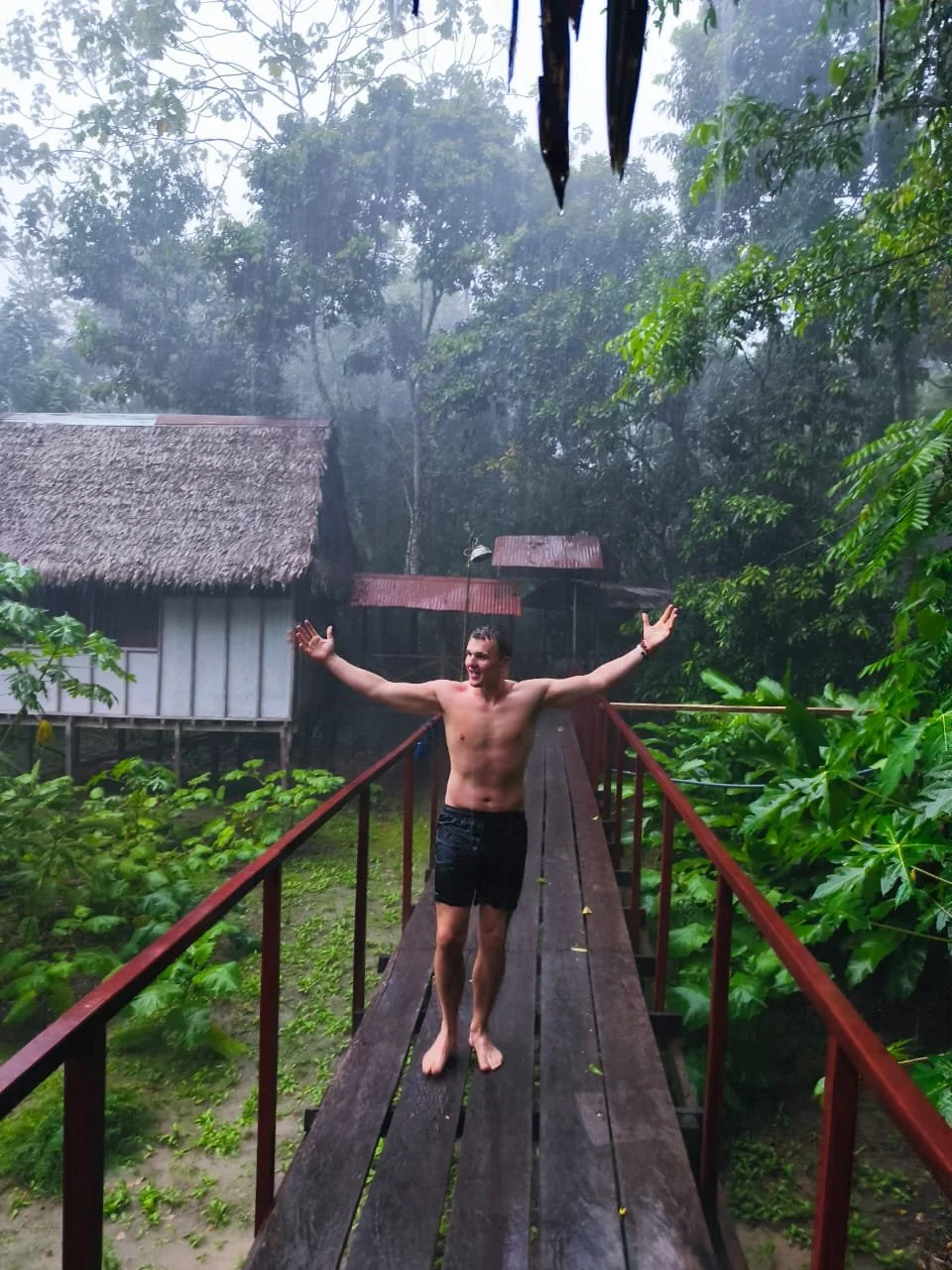 A man standing barefoot on a wooden bridge in a rainforest, with arms outstretched and smiling, surrounded by lush green foliage and a misty atmosphere.