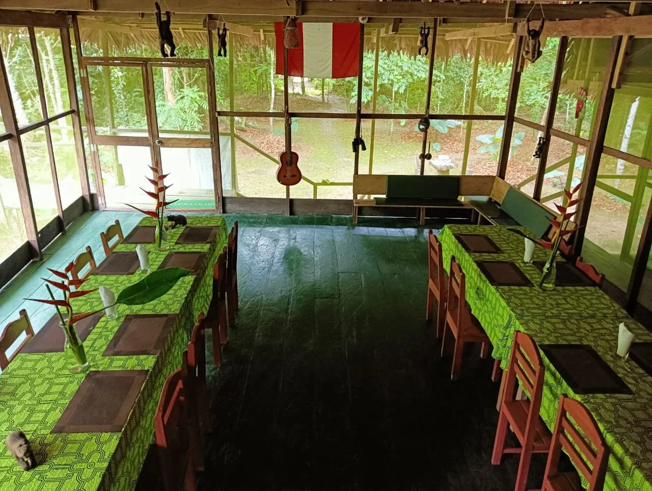 A rustic restaurant interior with two long tables covered with green patterned tablecloths, each set with brown placemats, white napkins, and vases with tropical flowers. The room has large windows overlooking a lush green outdoor area. Decorations include hanging black figurines and a guitar on the wall. The floor is dark wood or stone.