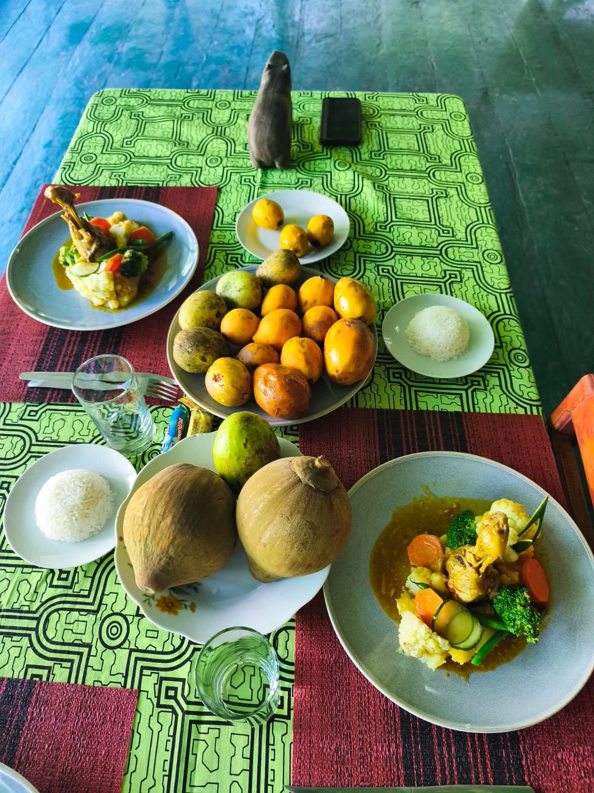 Table set with plates of rice, vegetable curry with chicken, various fruits including mangoes, bananas, and guavas, coconuts, and a glass of water, on a green and red patterned tablecloth.