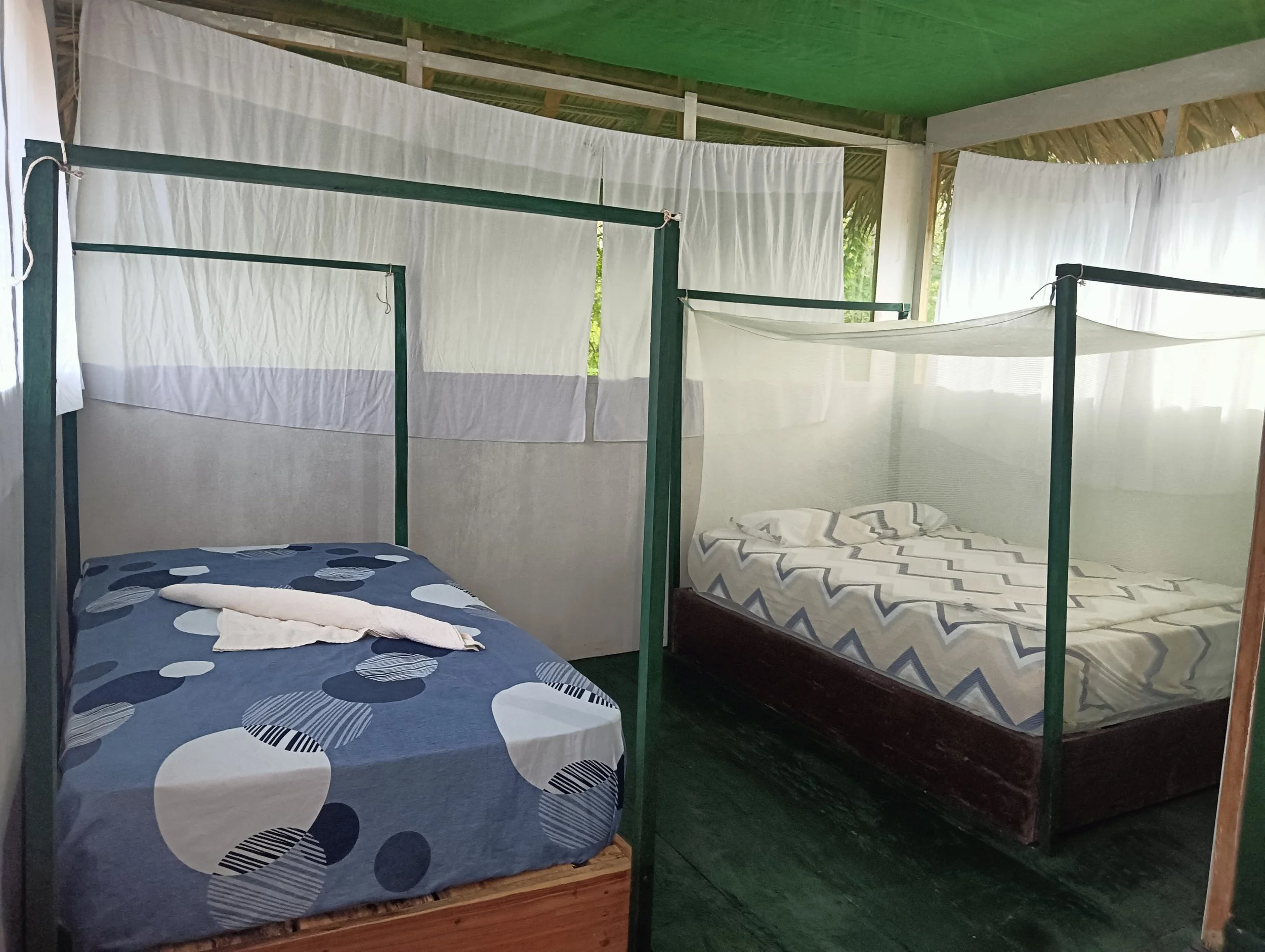 Two beds with mosquito nets in a tropical-style open room with green and thatched ceiling, and white curtains on the sides.