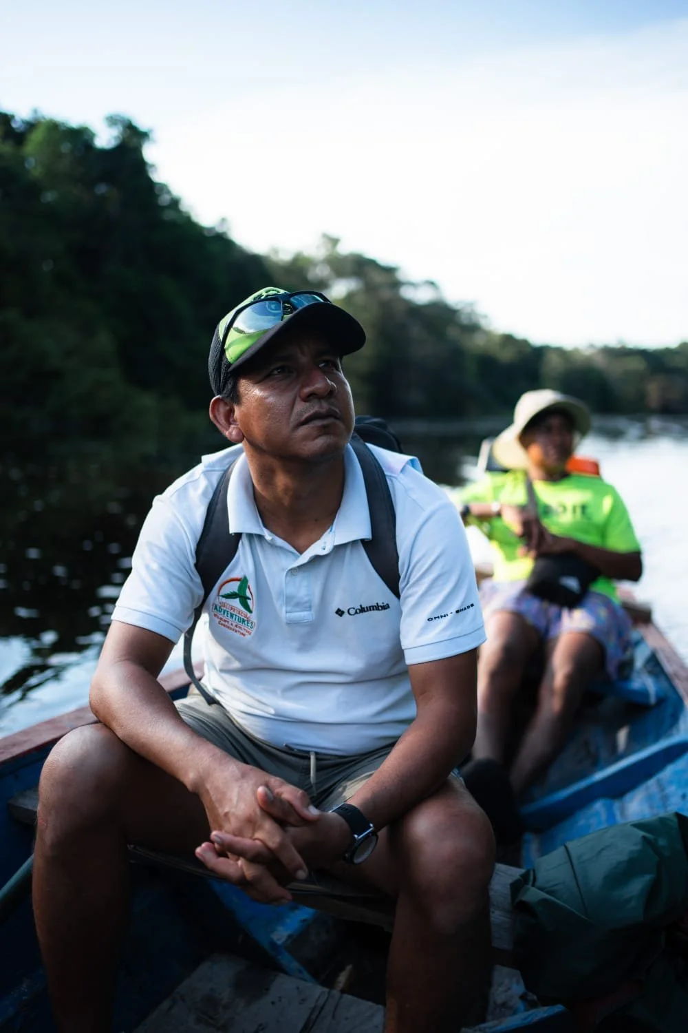 Two men in a boat on a river surrounded by trees, one in the foreground wearing a white polo shirt and a black cap with sunglasses, the other in the background wearing a bright green shirt and wide-brimmed hat.