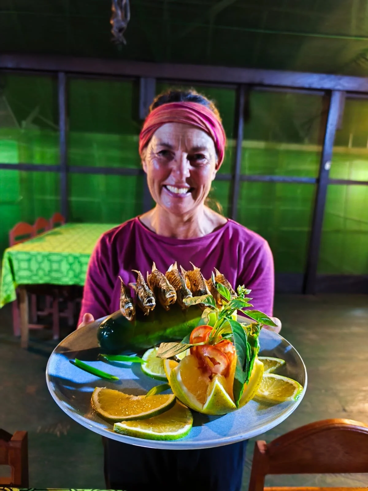 A woman with a pink headband holding a plate of traditional Mexican food, including rice, vegetables, and fried fish, with lime wedges on a restaurant table.