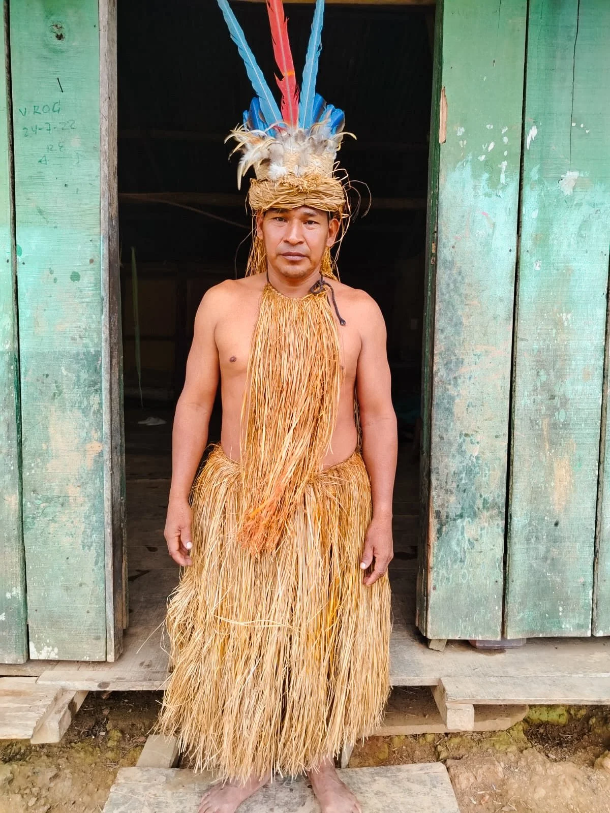 Man wearing traditional tribal attire, including a large feathered headdress, a grass skirt, and a grass necklace, standing in front of a wooden building.