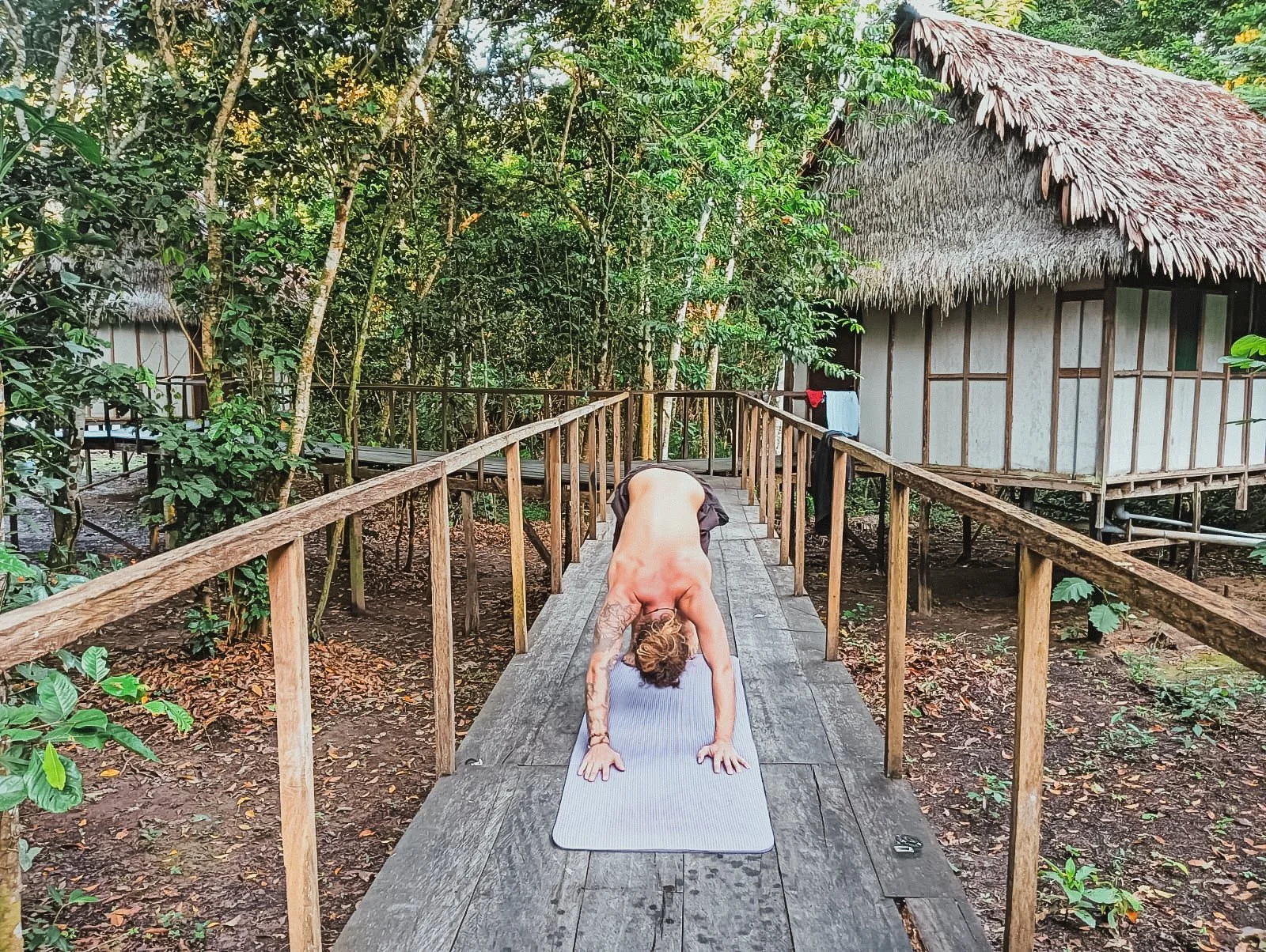 A person practicing yoga on a mat on a wooden bridge in a lush forest environment, with a rustic thatched hut visible in the background.