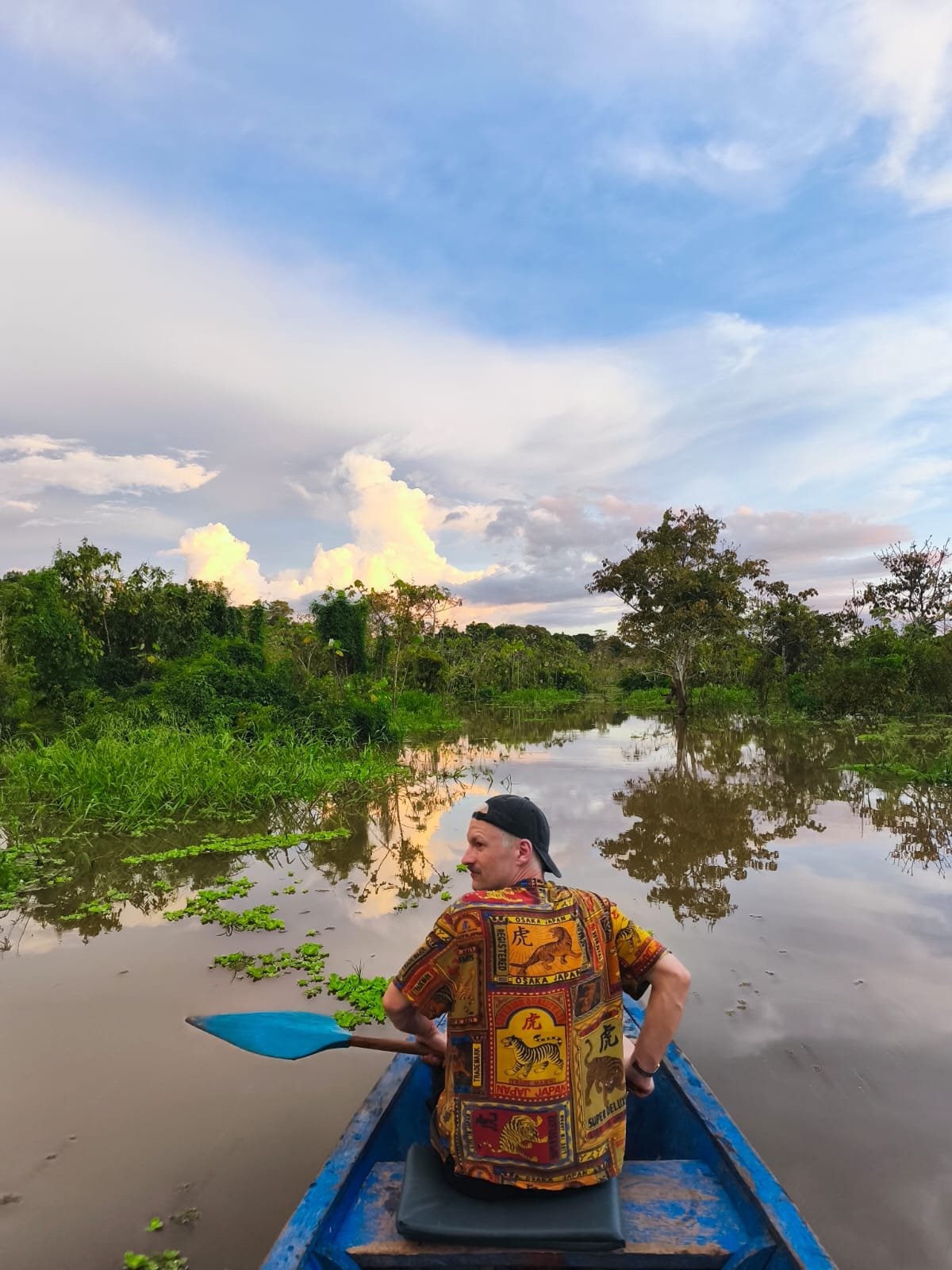 A man sitting in a small blue boat on a calm river, surrounded by lush green trees and vegetation, under a partly cloudy sky during sunset or sunrise.