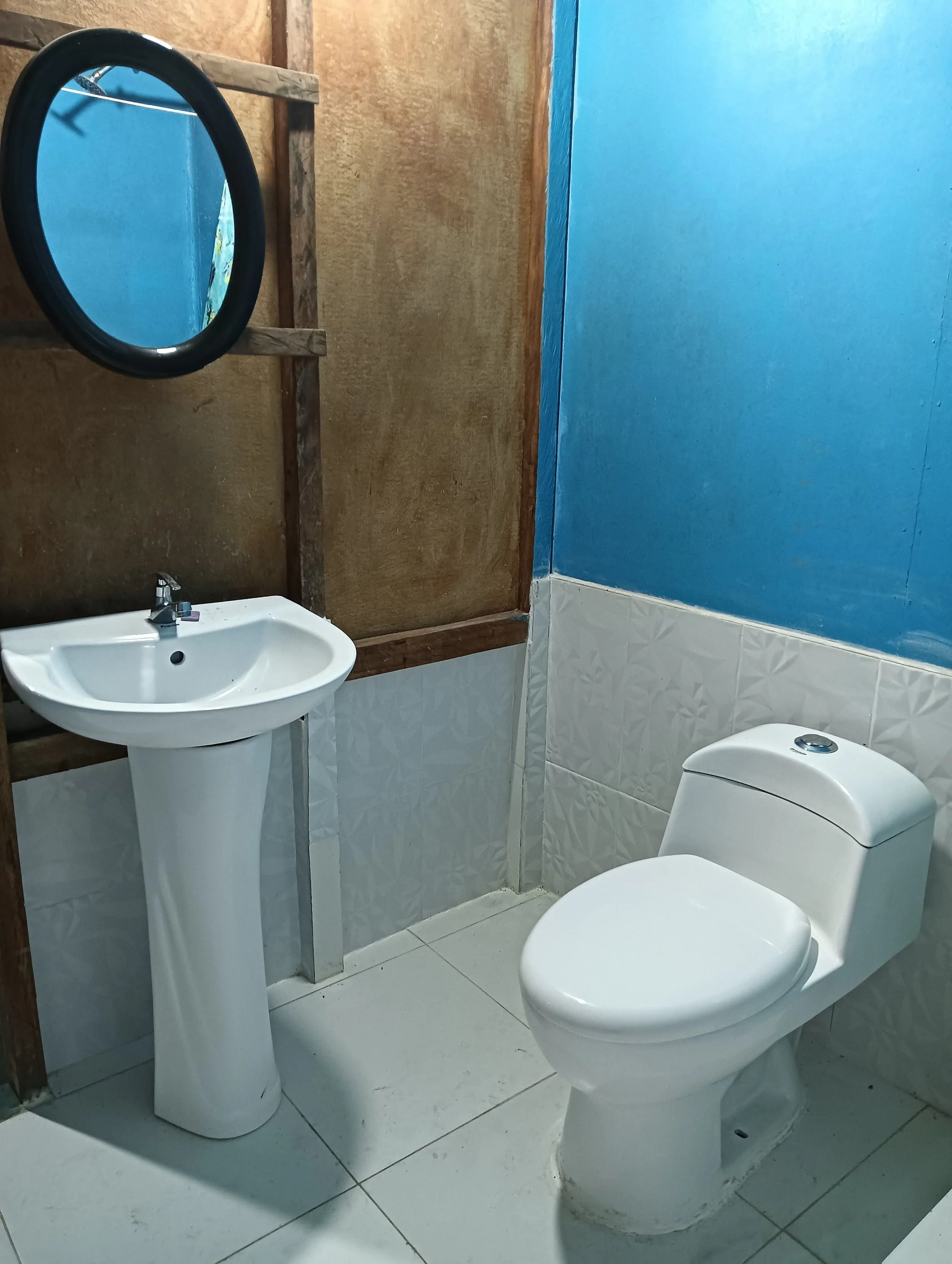 A small bathroom with a white toilet, a white pedestal sink, a round mirror, a blue wall, white textured tiles below, and unfinished wooden wall paneling.
