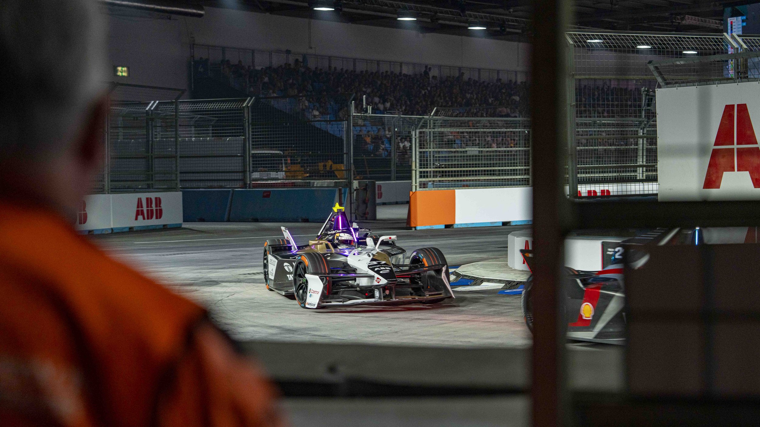 Nighttime race track with racing car after a crash, seen through a gate. Spectators in background.