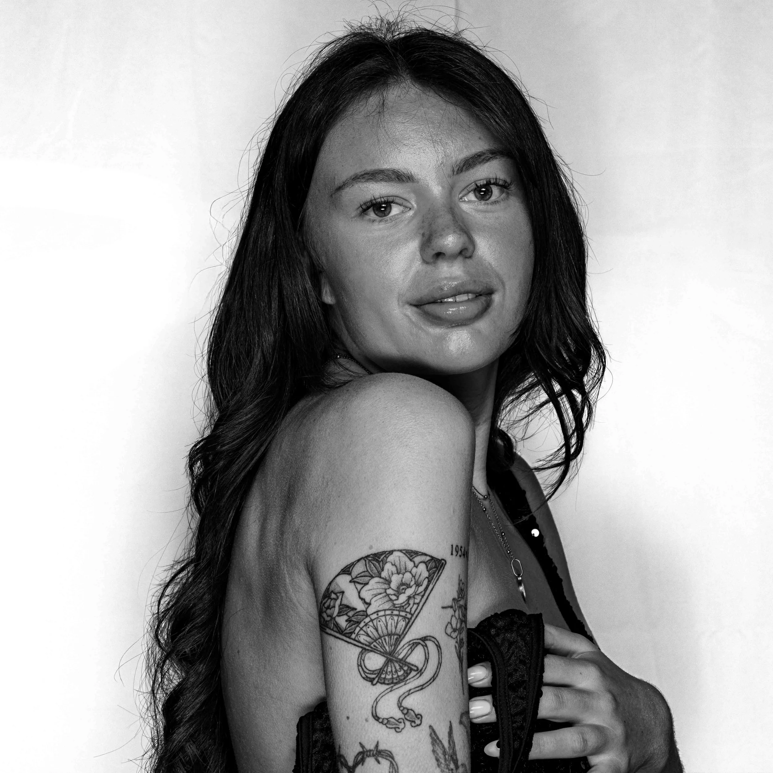 A woman with long dark hair, tattoos on her arm and chest, and a black leather vest, looking at the camera against a plain background.