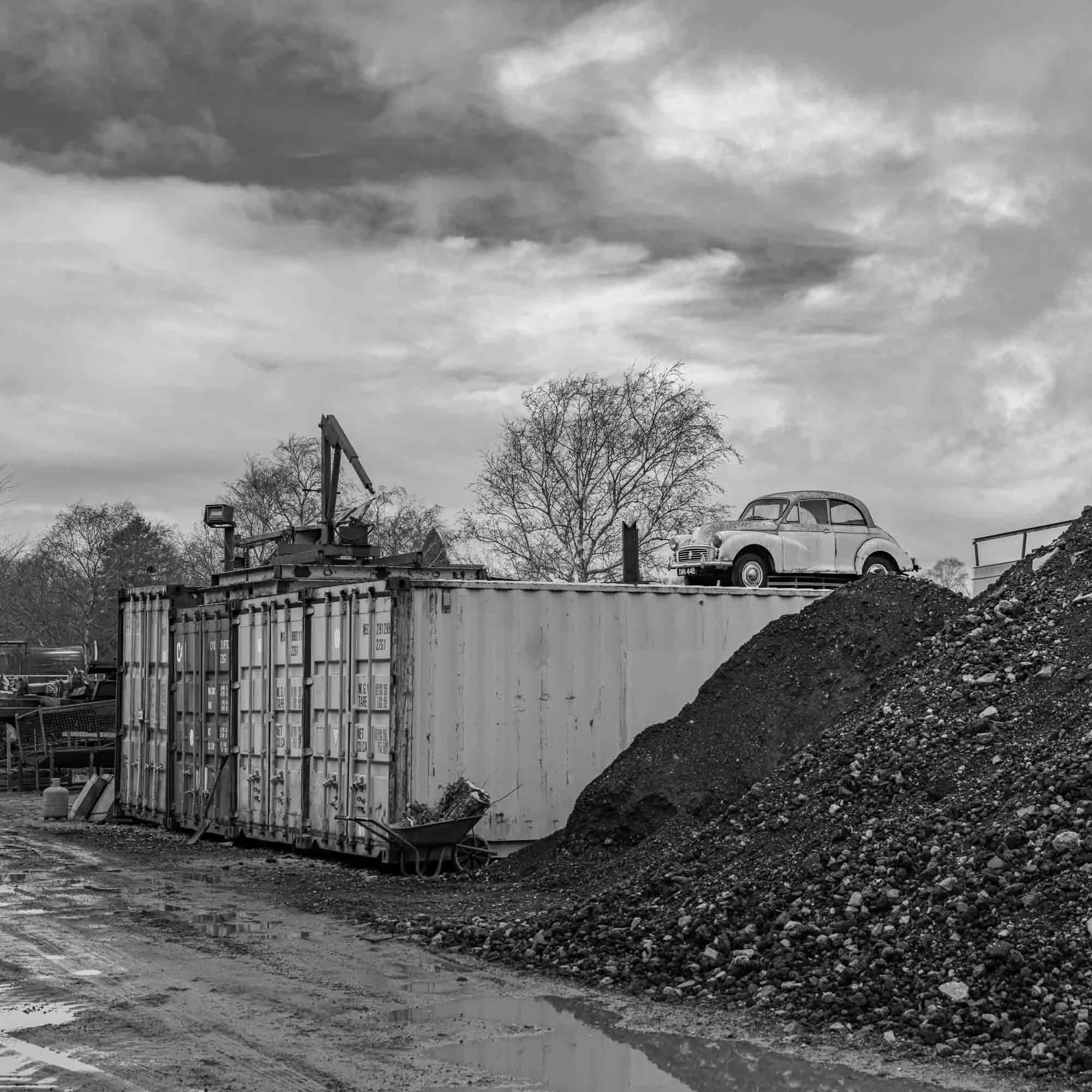 Black and white photo of a Morris Minor parked on top of a large shipping container, with a pile of dirt in the foreground and leafless trees in the background.