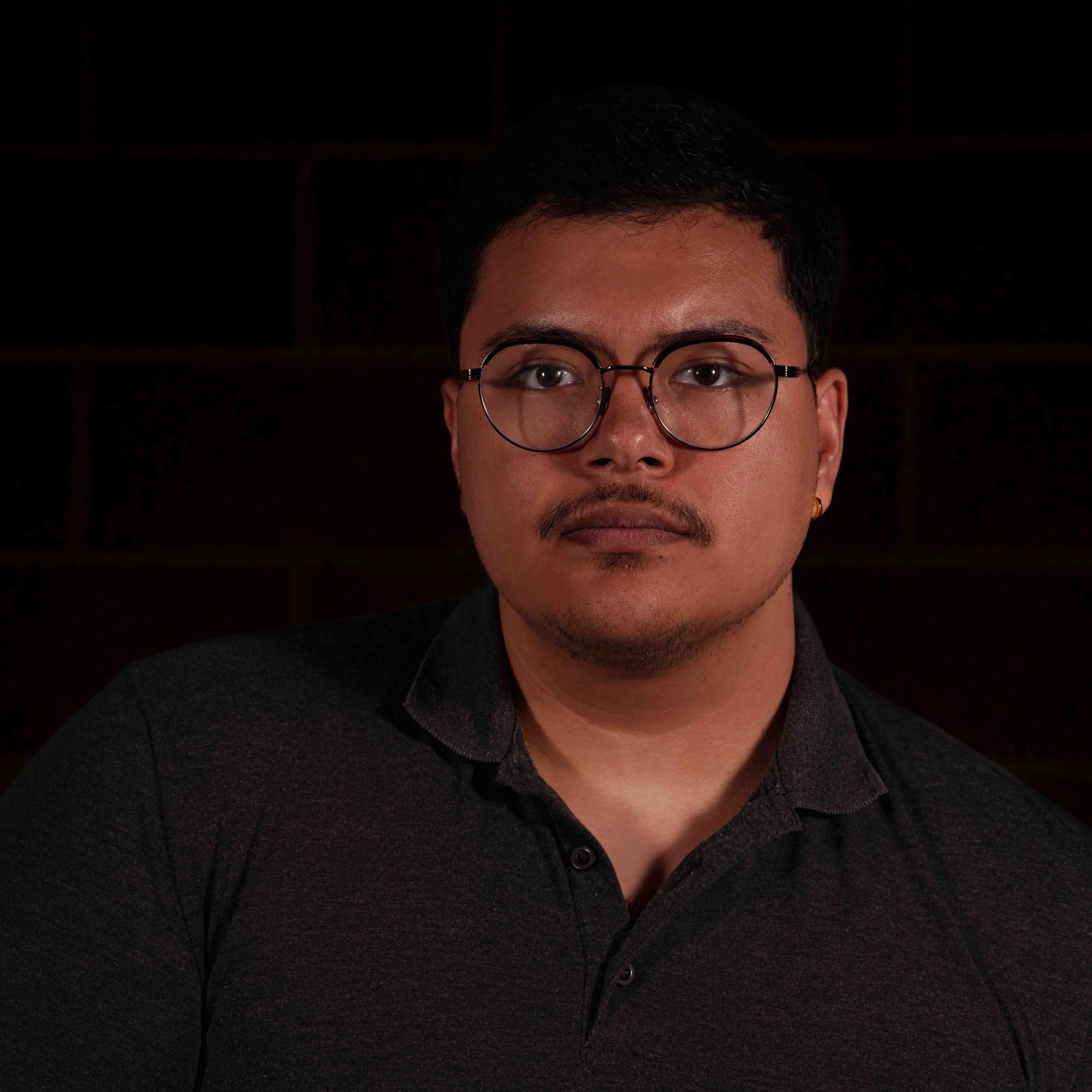 Close-up of a young man wearing round glasses and a black collared shirt, with a dark background.
