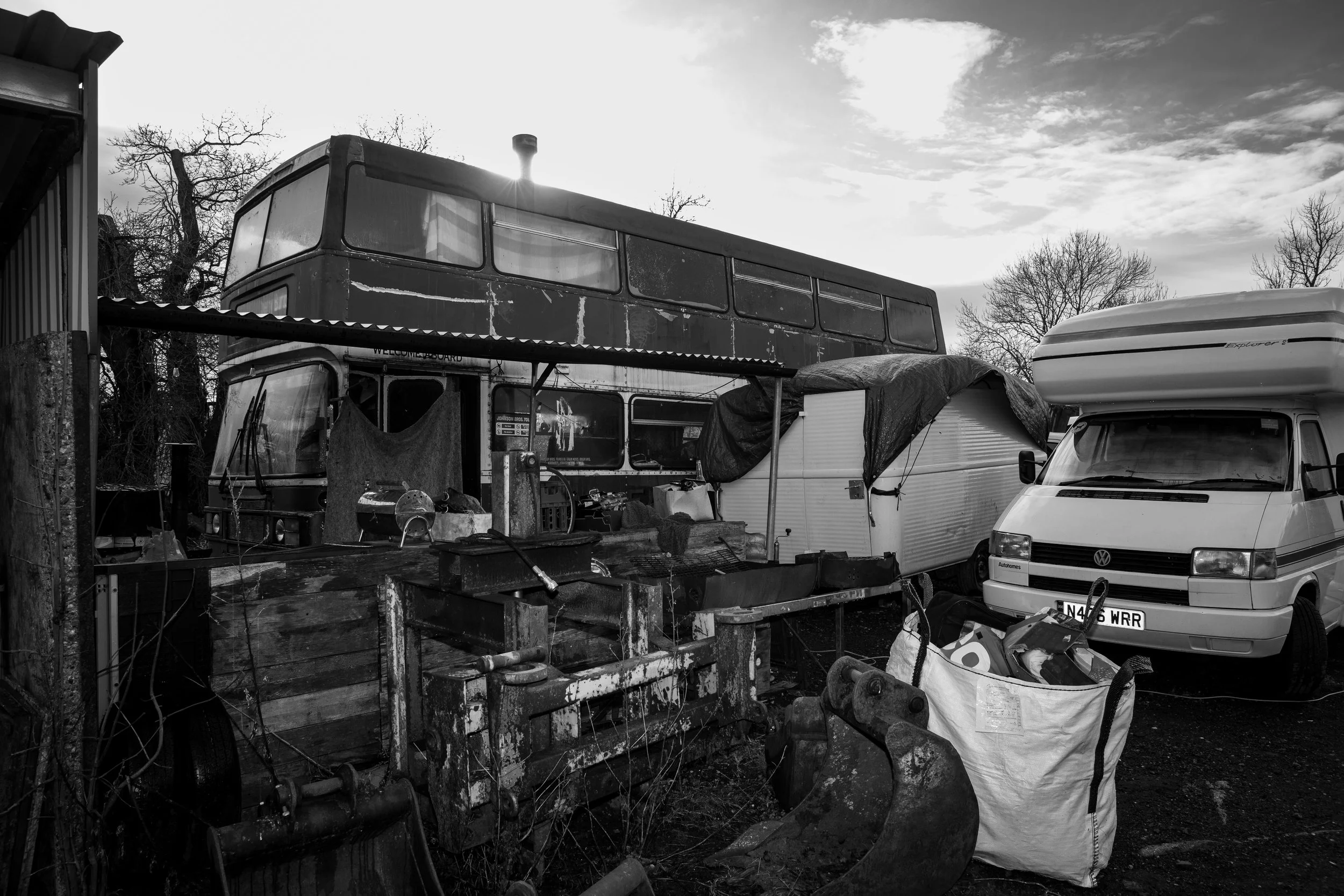 Black and white photo of an outdoor storage yard with old buses, trailer, pickup truck, and various tools and objects.