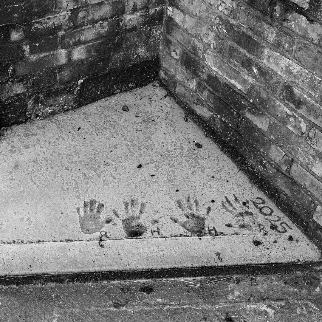 Two handprints and a single footprint pressed into concrete on a sidewalk, with some scattered dirt and debris. The handprints are marked with the letters 'C' and 'H', and the footprint with 'T'. There are also some dates and other markings near the 