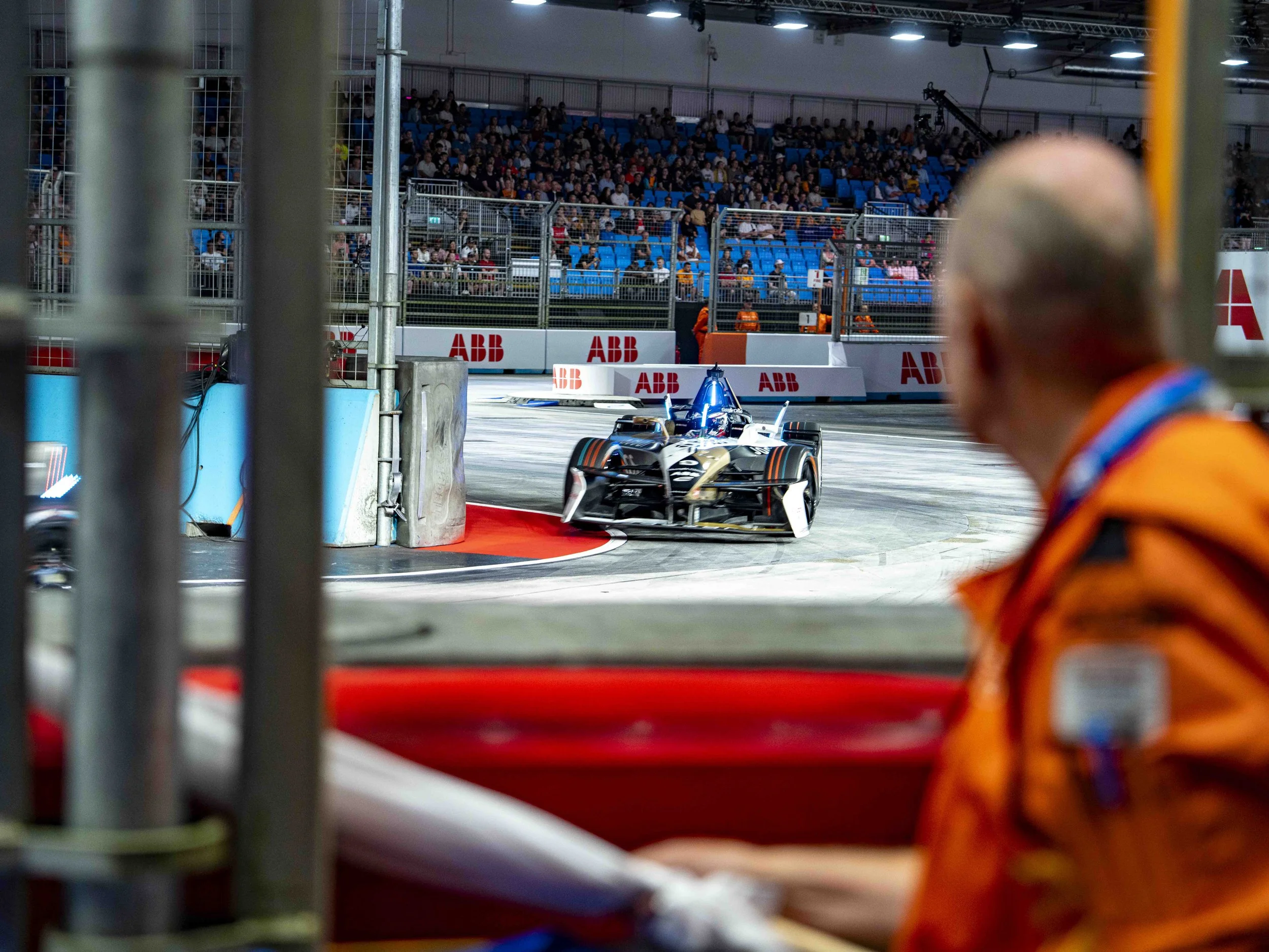 Race car on a track viewed from behind a barrier, with a Marshall in the foreground, and a crowd in the stands in the background.